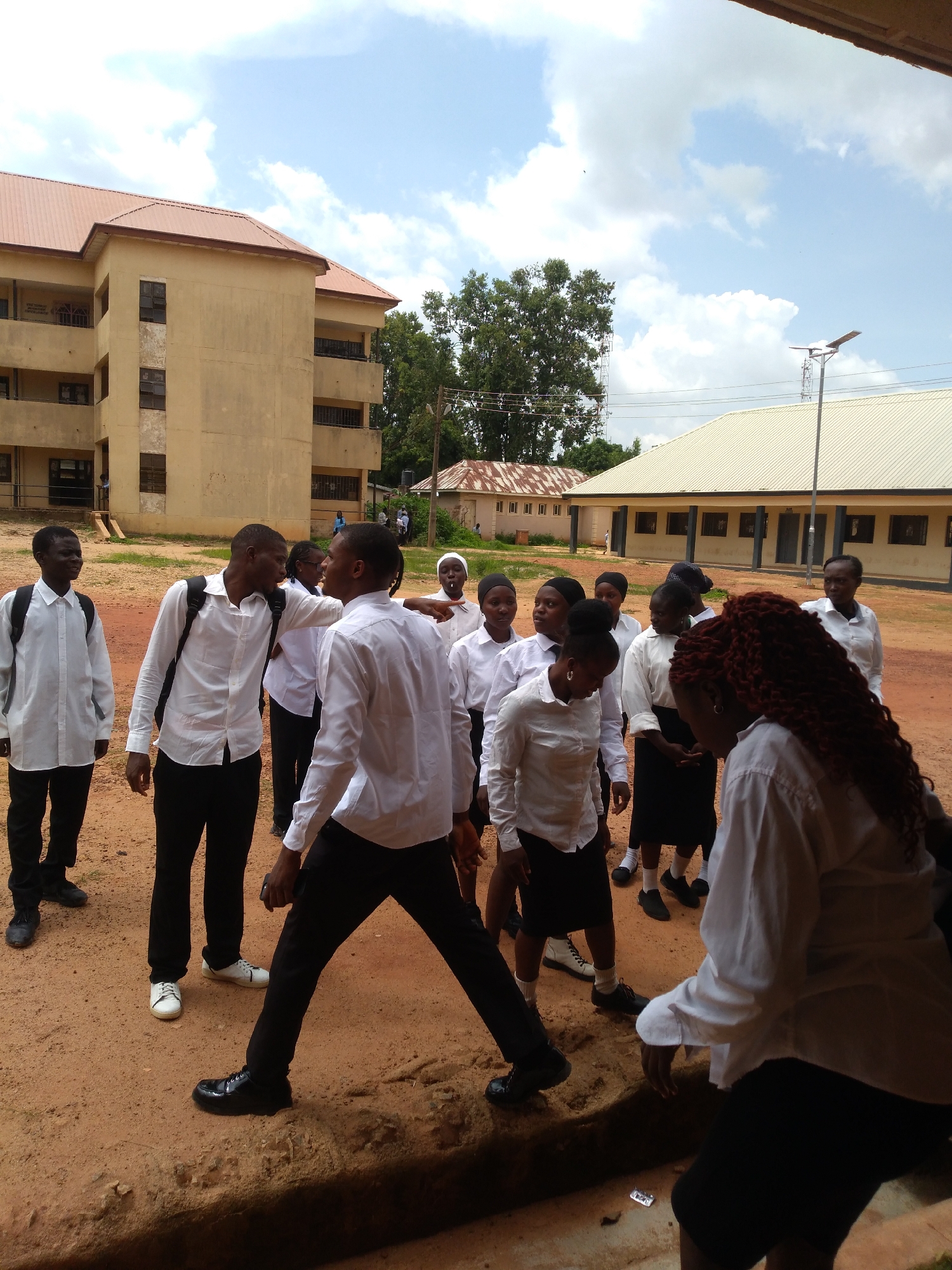 In the city of Jos, Nigeria, a group of young people in white shirts and black trousers or skirts are gathered outdoors near a school building. The building, a multi-story structure with a red roof, appears to be made of beige-colored material. A smaller building with a white roof and a tall pole with a solar lamp is also visible in the background. The ground is covered in reddish-brown dirt. Some individuals are interacting, with one person gesturing towards another. The sky is partly cloudy.