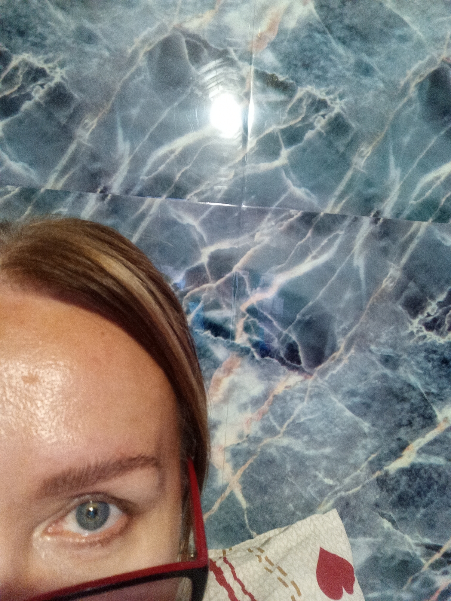 The image is a close-up, slightly tilted shot of a woman's face and upper body, taken in Novopokrovka, Russia. Her left eye, framed by dark eyebrows and red glasses, is the focal point. The background consists of a marble-patterned wall, with a bright light source reflecting near the top. A portion of patterned fabric, possibly clothing or bedding, is visible in the lower right corner.