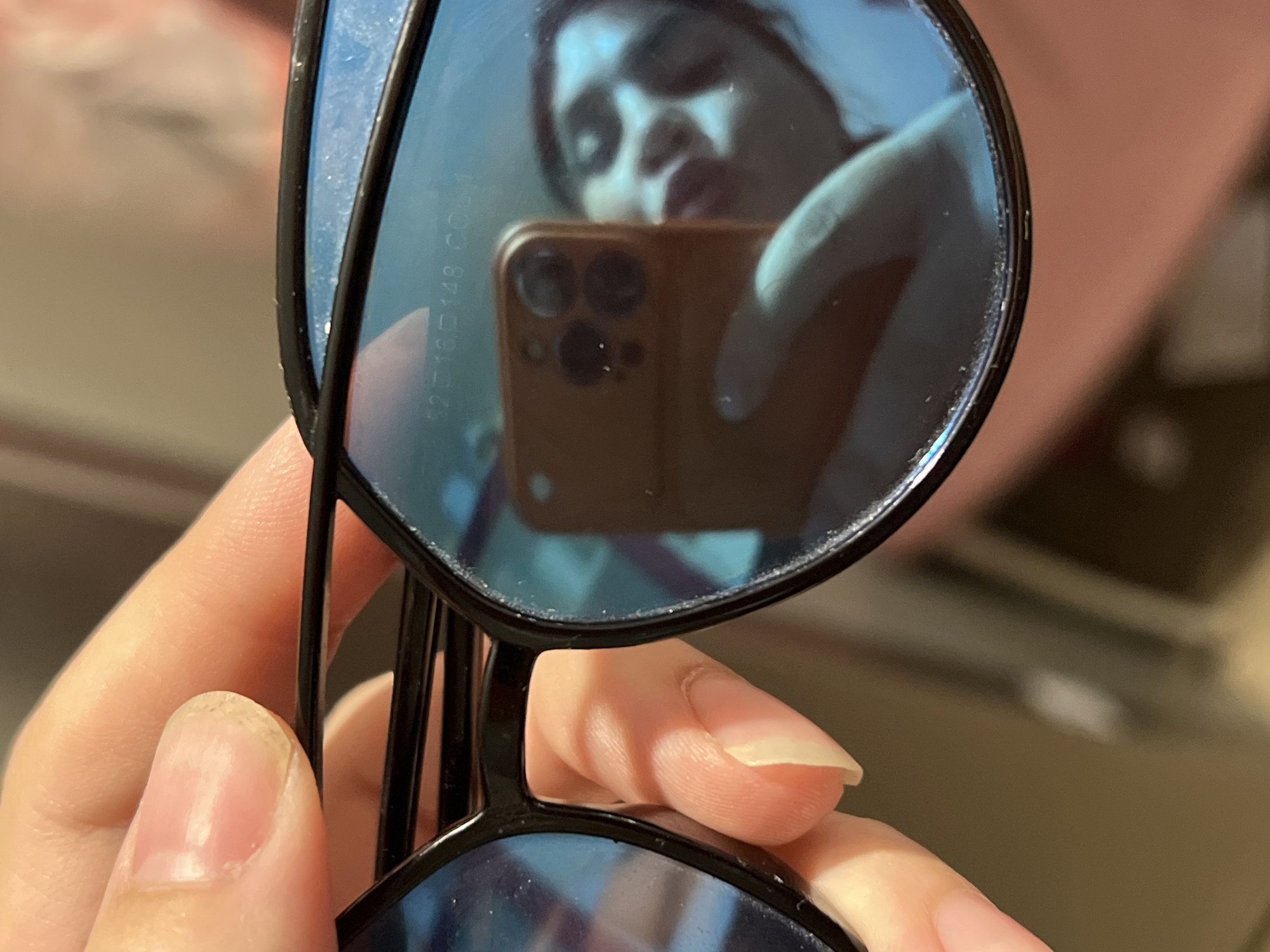 A person holds a pair of sunglasses, reflecting a selfie in its lenses. The reflection shows the person holding a phone, presumably taking the picture. The environment is a room with hints of furniture and personal items, but is not definitively located in the city of Noviy, Russia.