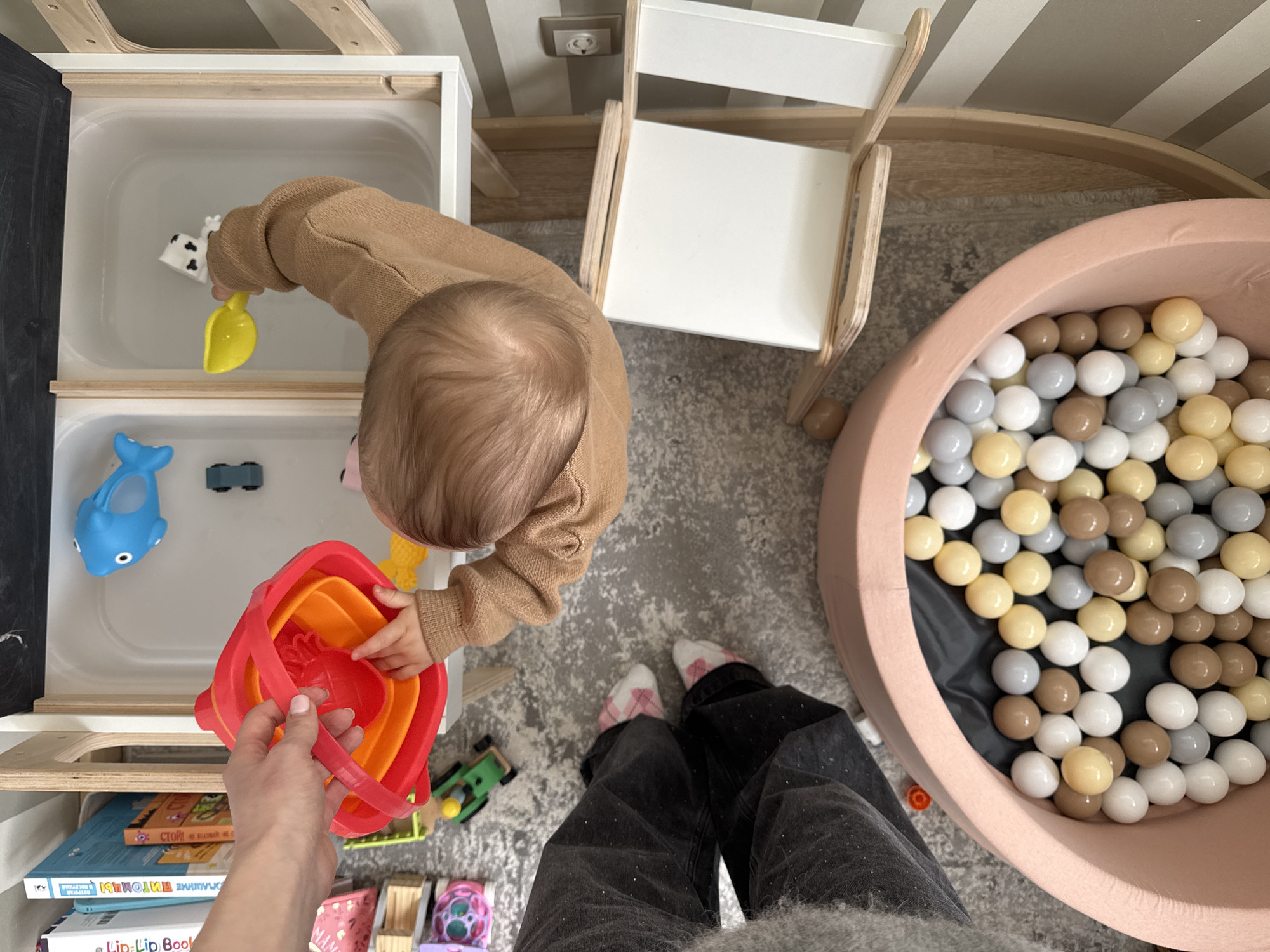 An overhead shot shows an indoor scene, likely a playroom, with a baby standing near a toy storage unit. The baby, wearing a tan sweater, is reaching into a stack of colorful buckets held by an adult. The storage unit, on the left, contains toys, including a blue dolphin and a black block, as well as books on the lower shelves. Adjacent to the baby is a small white chair with a wooden frame. To the right is a round, pink ball pit filled with white, gray, and tan balls. The floor is gray with a subtle pattern. The adult is wearing dark jeans with light-colored socks.
The location is likely Moscow, Russia, due to the presence of Russian language books on the storage unit shelves.