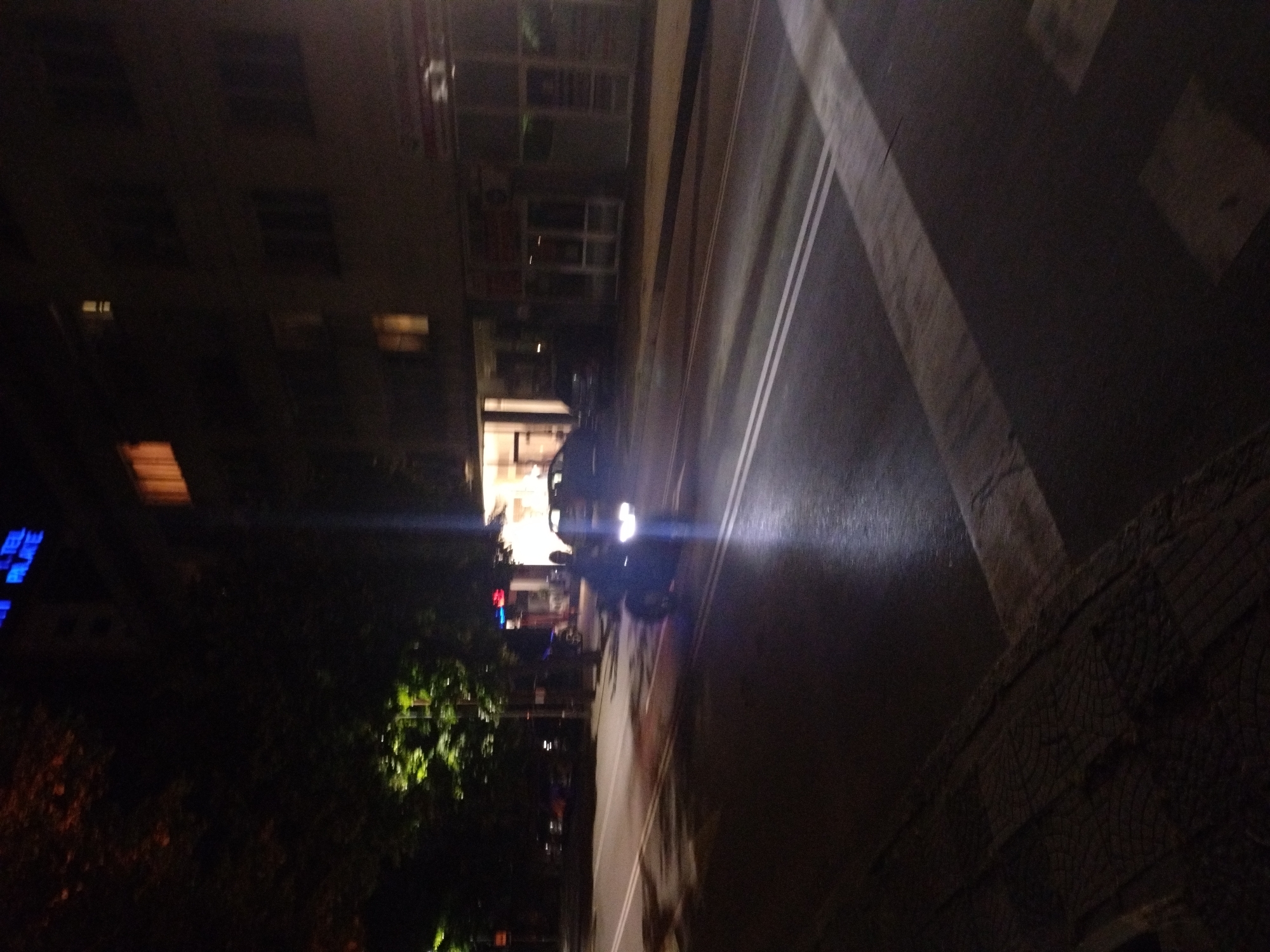 The image shows a nighttime scene in Stara Zagora, Bulgaria. A wet road with double white lines runs diagonally across the frame. A dark car with headlights on is driving on the road. To the left of the road is a building with lit windows and signage, partially obscured by trees. The environment is dimly lit, suggesting nighttime. There's a sidewalk area visible on the right, with the texture of paving stones. The buildings, cars and trees suggest an urban environment.