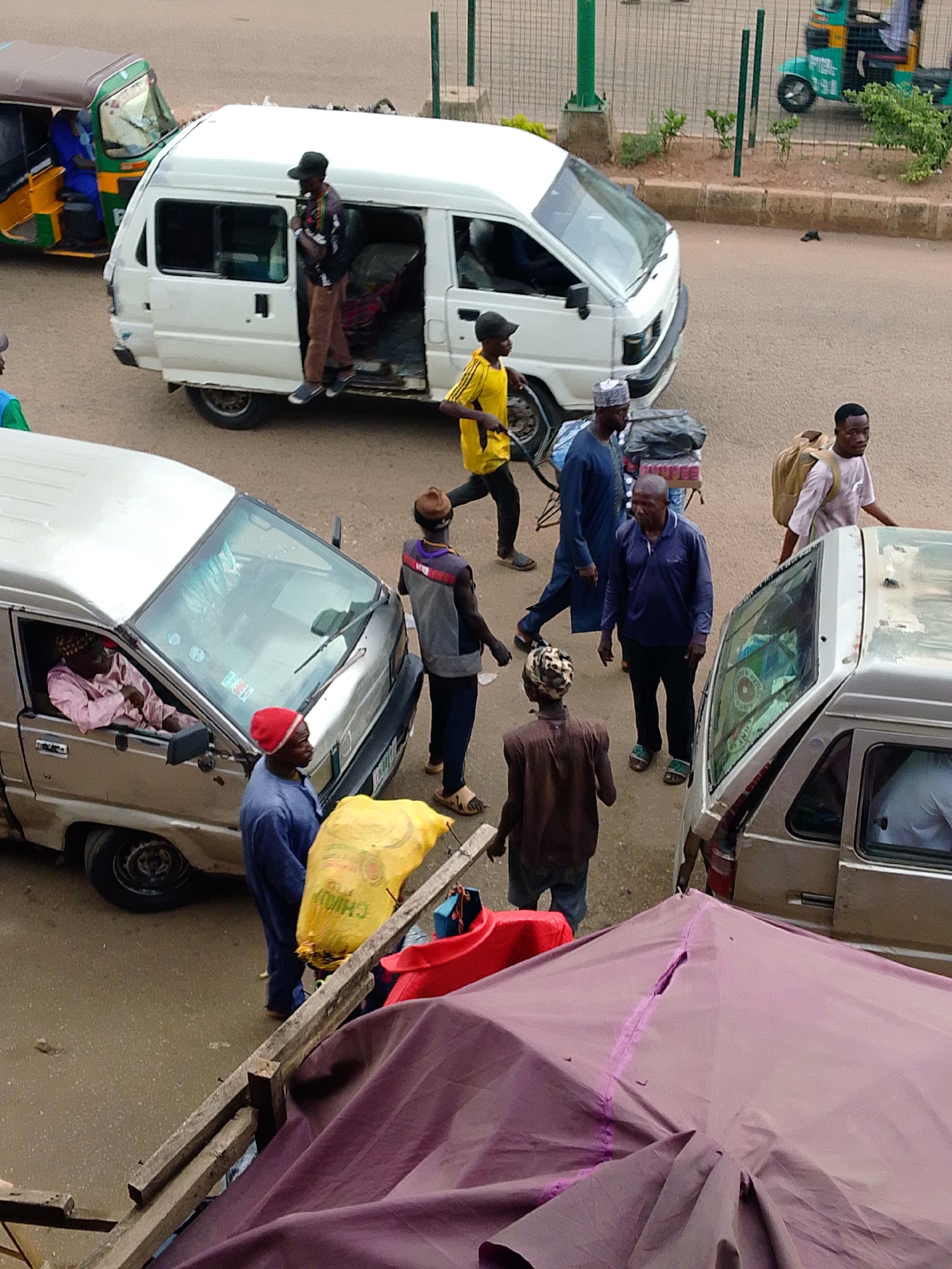 The image depicts a street scene in Bukuru, Nigeria, featuring multiple individuals and vehicles. A white minibus is parked on the left, with its door open and several people standing around it. One person is partially inside the minibus, and another is stepping out. Adjacent to the minibus, a person in a yellow shirt walks away from the camera. To the right of the white minibus, a silver minivan is parked. A woman is visible inside the silver minivan through the driver's side window. A man wearing a red hat stands next to the silver minivan, holding a large yellow bag. Further to the right, another man stands with a backpack. In the foreground, a large purple tarpaulin is draped over a structure, partially obscuring the view. The street is paved, and a green fence is visible in the background. The overall impression is of a busy public space, possibly a transport hub or market area.