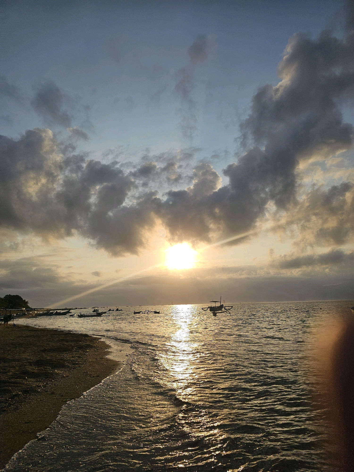 This image depicts a serene beach scene at sunset in Denpasar, Indonesia. The sky is filled with dramatic clouds, with the sun peeking through, casting a golden glow on the ocean. The water is calm, with gentle waves lapping at the shore. Several traditional Balinese fishing boats, known as jukung, are anchored offshore, their silhouettes adding to the picturesque view. The beach itself is made of dark sand, with some vegetation visible on the left side of the frame. The overall atmosphere is one of tranquility and natural beauty.