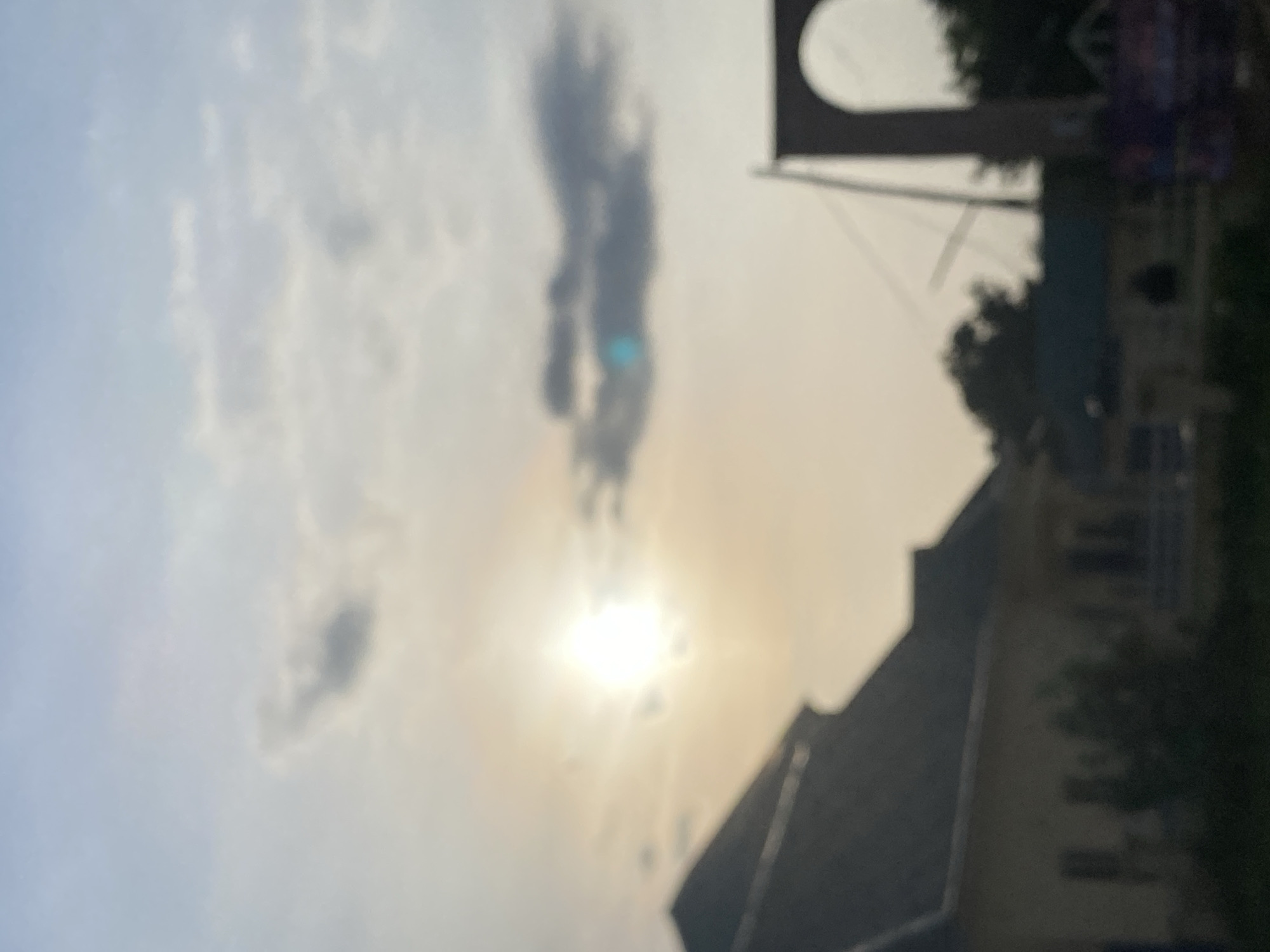 The image depicts a blurry, outdoor scene at sunset or sunrise, likely in Jalingo, Nigeria. In the foreground, a large, dark-roofed building is partially visible on the left. To the right, an archway structure, possibly part of a bridge or monument, is present. Trees and other vegetation are scattered throughout the background, some appearing dense. The sky is filled with a soft, hazy light, with the sun appearing as a bright, diffused orb, casting a warm glow across the scene. Clouds are present, some appearing dark and others lighter, suggesting a dynamic sky. The overall impression is one of an urban or semi-urban environment under a diffused, ambient light, typical of twilight.