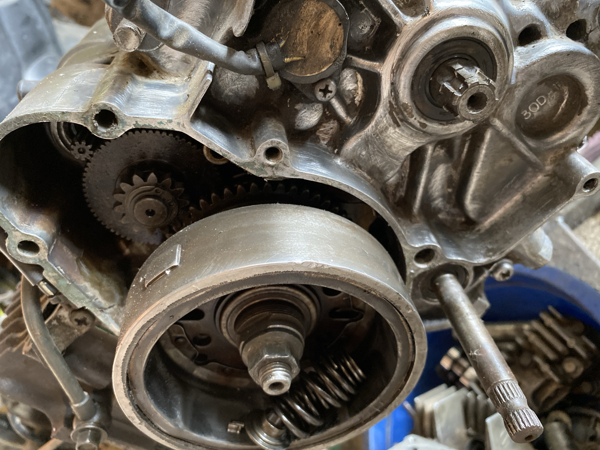 The image shows a close-up of a disassembled motorcycle engine, with a focus on the flywheel and internal gears. The engine casing appears to be made of aluminum and shows signs of wear and tear, with some dirt and grime visible. Several gears are visible within the casing, intermeshing with each other. A large, circular component, likely the flywheel, is prominent in the foreground, with springs and a central nut visible. A metal shaft with splines is also visible extending from the engine. The background is out of focus but suggests a cluttered environment, possibly a workshop or garage. There are no people or animals present in the image.