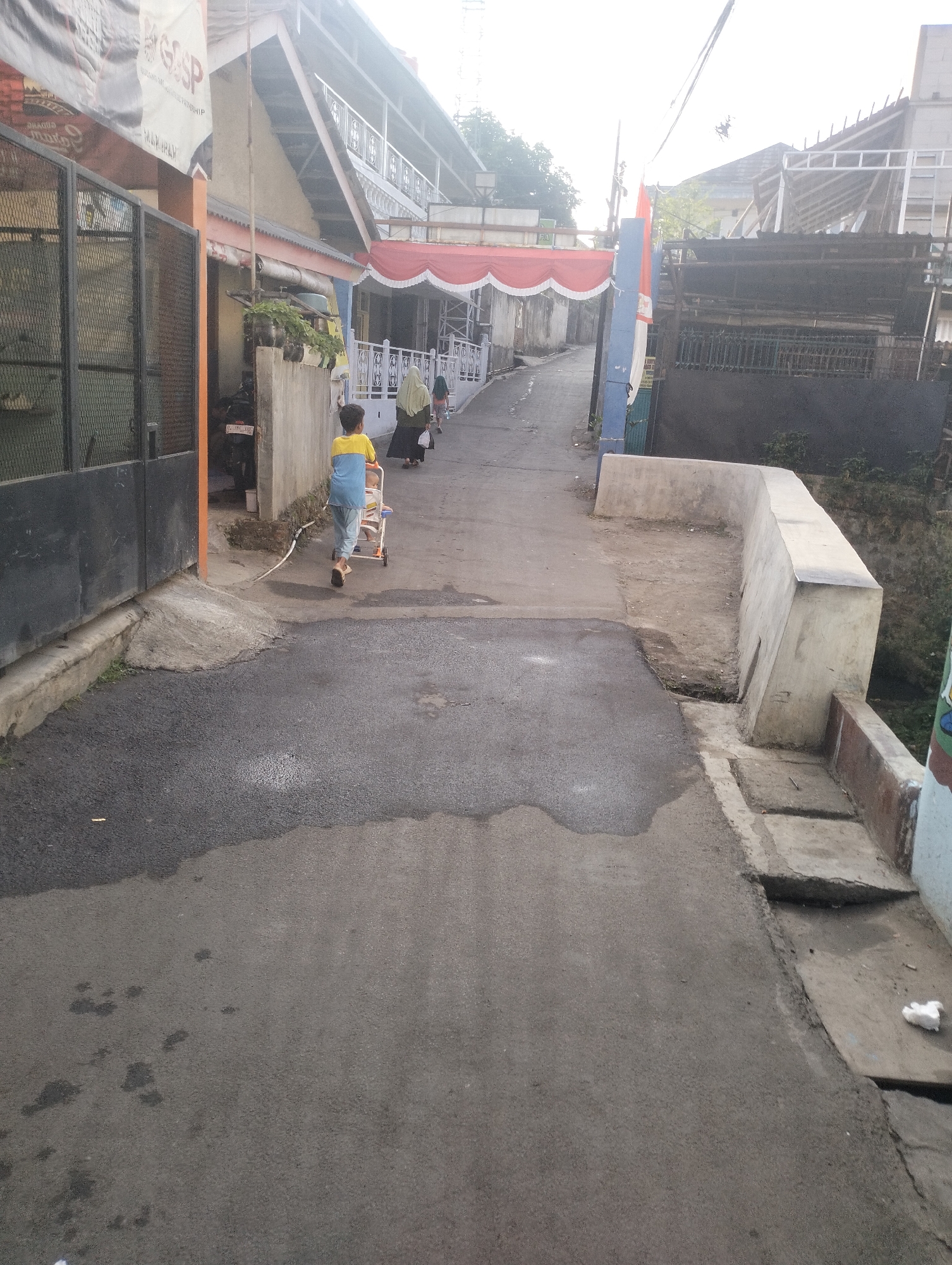 A young person wearing a blue and yellow shirt and light-colored pants is walking away from the camera, pushing a small, multi-colored stroller along a paved street. The street is lined with buildings and fences. A red and white banner is stretched across the street in the background, and an Indonesian flag is visible on a pole. The area appears to be in a residential or commercial district, with signs of ongoing construction or renovation in some buildings. The environment is outdoors, with natural lighting suggesting daytime. The overall impression is a street scene in Indonesia.