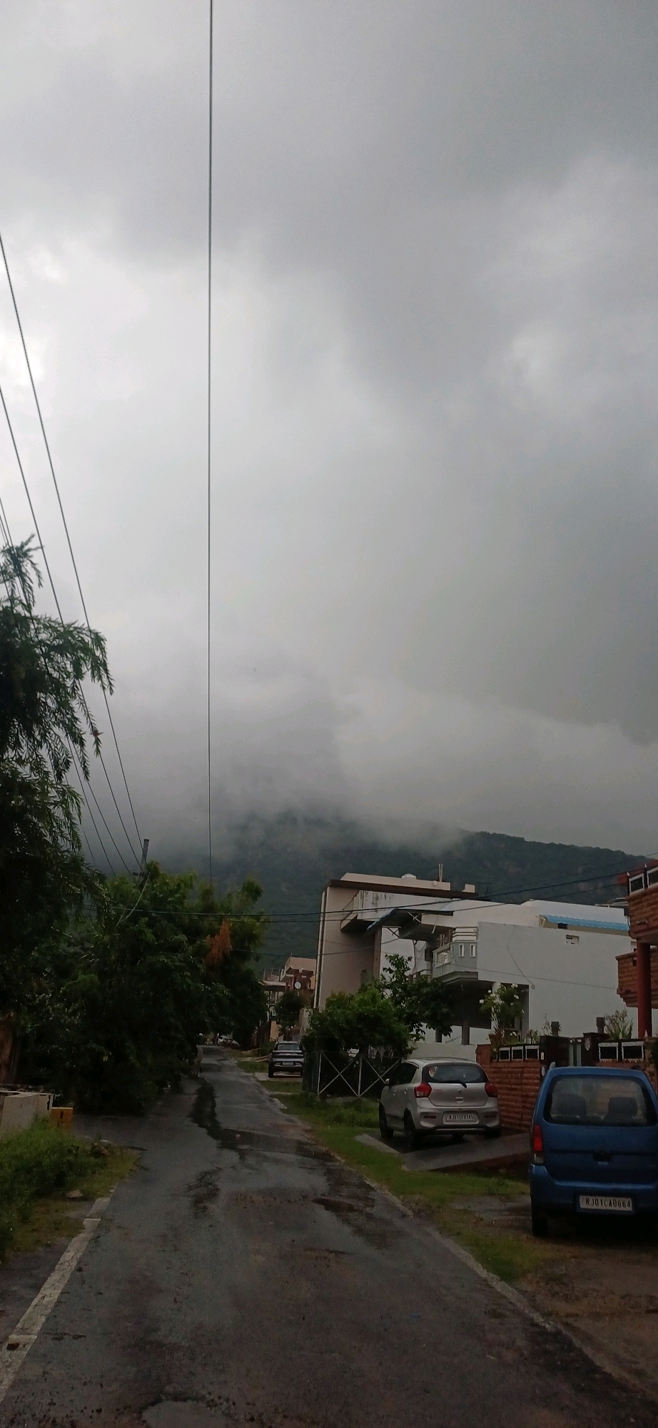 The image depicts a street scene in the city of Ajmer, India, under a heavily overcast sky, suggesting an impending or recent rain. The road is wet and appears to have some potholes, indicating it is a typical urban or suburban street. On the left side of the road, lush green trees and foliage line the street, with power lines running diagonally overhead. Several modern houses with white and beige facades are visible on the right side of the road. Two cars are parked on the right side of the street: a silver hatchback and a blue van. In the background, a misty mountain range is visible, partially obscured by clouds. The overall atmosphere is somber and damp.