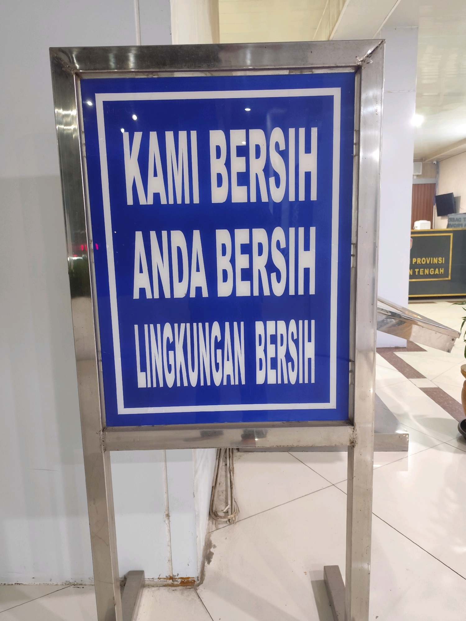 A blue and white sign on a silver metal stand reads "KAMI BERSIH ANDA BERSIH LINGKUNGAN BERSIH". This translates to "We are clean, you are clean, the environment is clean". In the background, a counter is visible with the words "PROVINSI N. TENGAH" which likely refers to the province of Central Kalimantan, Indonesia. This suggests the location of the sign is within a public building or office in Palangka Raya, the capital city of Central Kalimantan. The floor is tiled with a light-colored pattern, and a white wall is visible to the left. Some electrical wiring is seen bundled along the floor and wall in the lower right portion of the image.