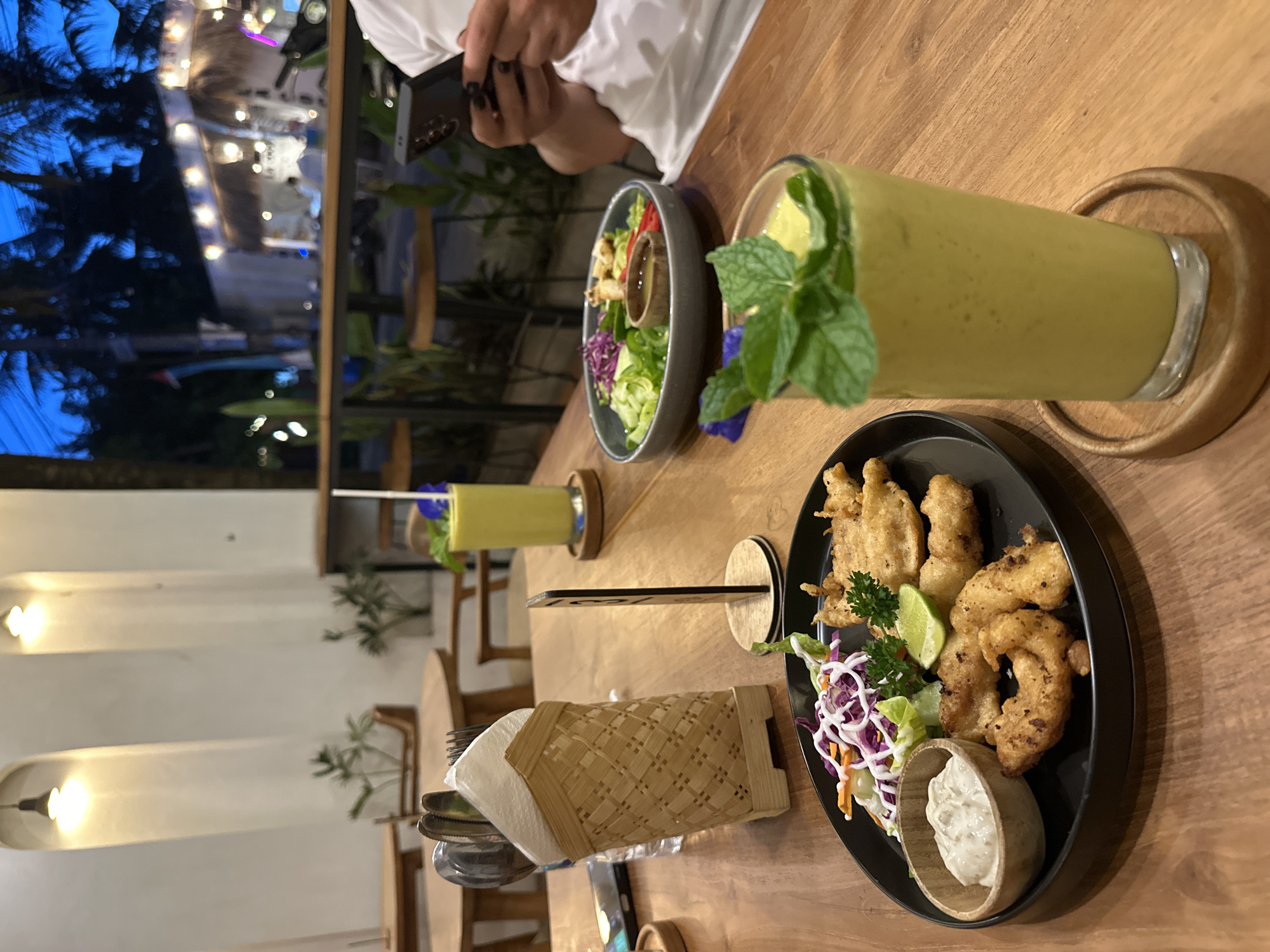 The image shows a wooden table in a restaurant in Nusa Penida, Indonesia. On the table, there is a black plate with fried food, a small salad, and a bowl of sauce. Next to the plate, there is a woven basket containing silverware and napkins. A glass of green smoothie with mint leaves on top is placed on a wooden coaster. In the background, there is another glass of green smoothie and a bowl of salad. A person is visible in the background using a phone. The background shows the restaurant interior and an outdoor scene with lights and plants. The atmosphere appears to be relaxed and the lighting is soft.