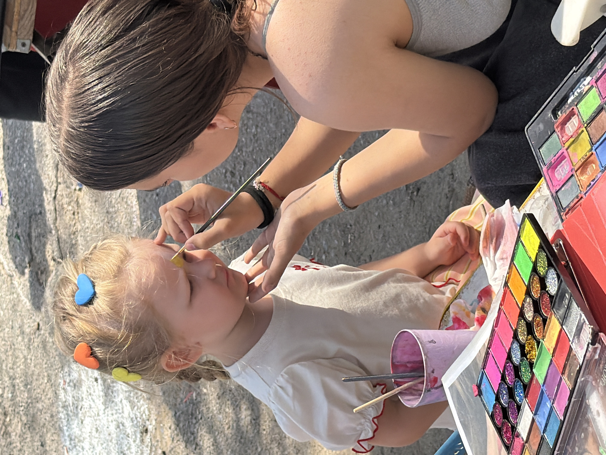 A woman is applying face paint to a young child. The child is fair-skinned and has light blonde hair with colorful heart-shaped hair clips. She is wearing a white shirt with red trim. The woman has dark hair and is wearing a gray tank top. She is using a brush to apply yellow paint to the child's forehead. Face paint palettes are visible, suggesting a street fair or similar event. The setting appears to be outdoors, with a paved surface visible. No information about the city of Kamyshin, Russia is discernable in the image.