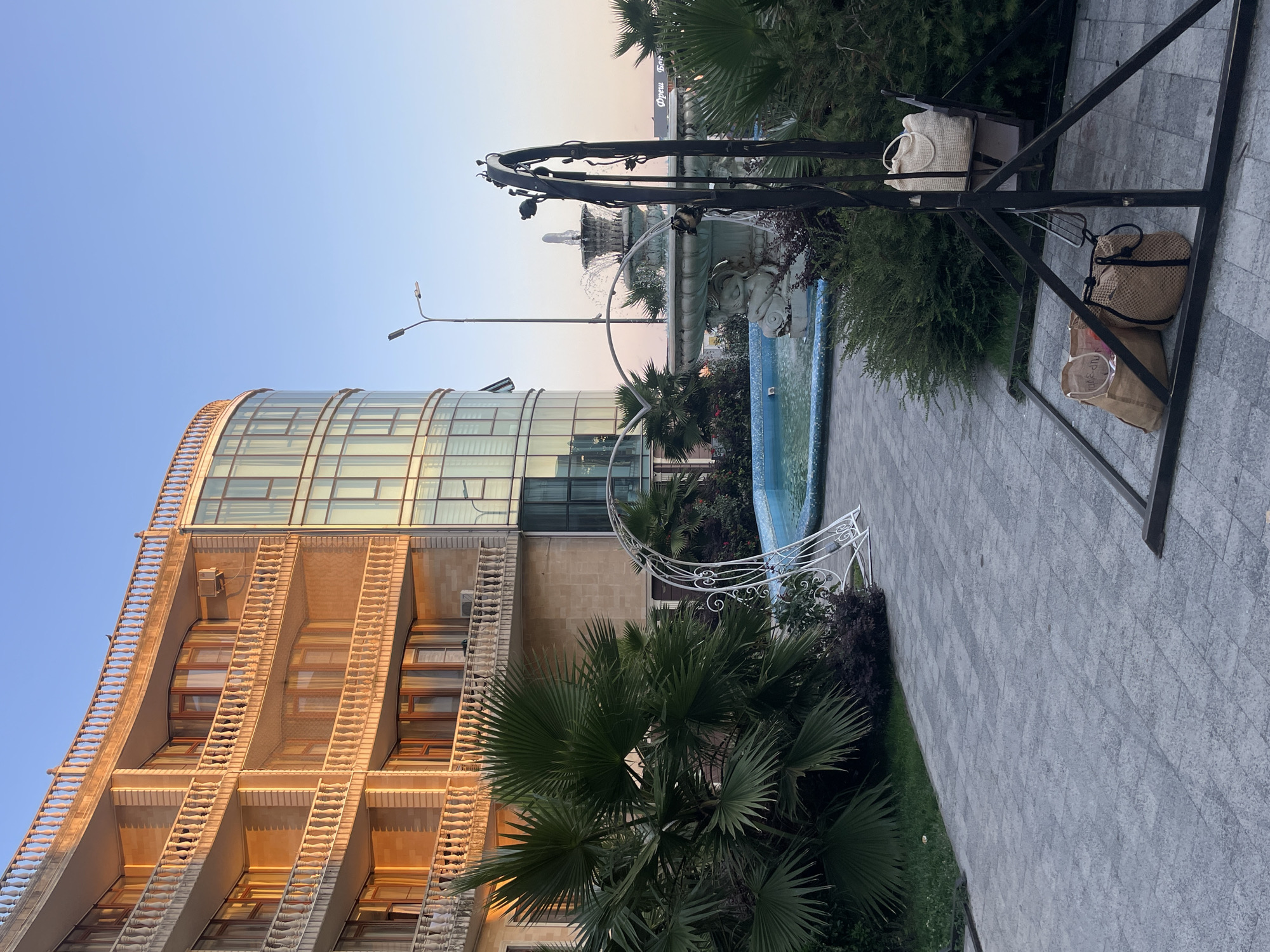 The image depicts a sunny outdoor scene in Sochi, Russia, featuring a modern building with a curved facade and balconies. In the foreground, a paved pathway leads to a decorative fountain with water jets and sculpted figures. Lush green palm trees and other foliage surround the fountain and the pathway. A metal swing set with two bags hanging from it is also present in the foreground. The sky is clear and blue, indicating a pleasant day. The lighting suggests it might be late afternoon or early morning, with the sun casting a warm glow on the building.