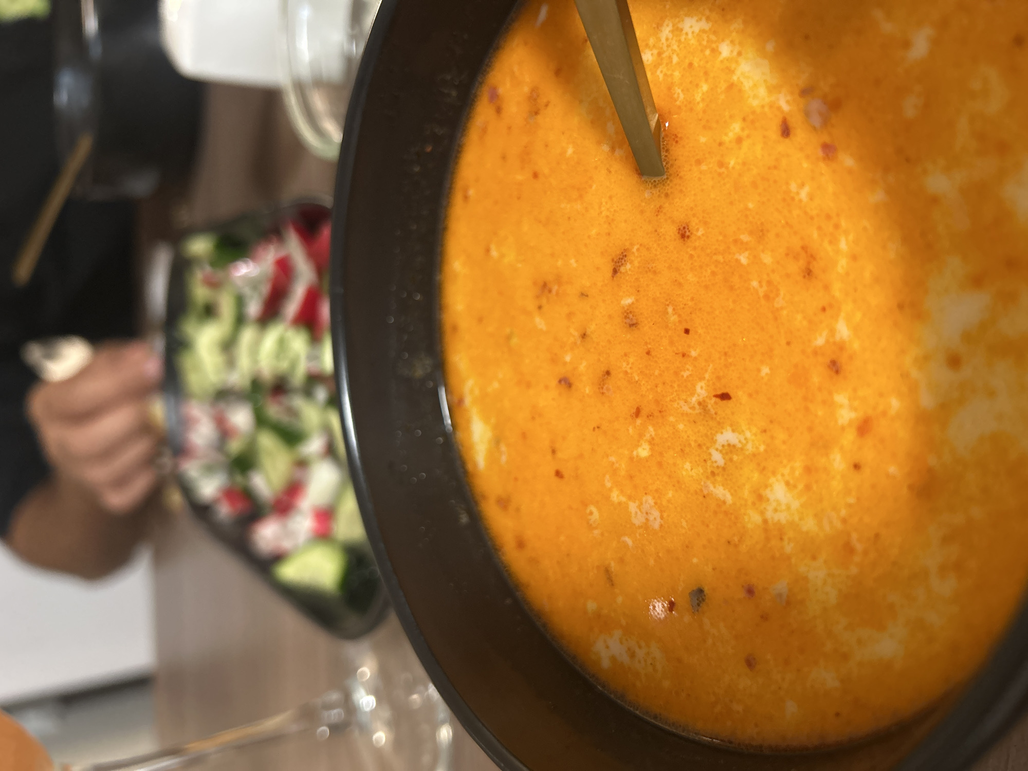 A close-up, angled shot shows a bowl of orange soup with a spoon in it, next to a salad with cucumbers and radishes. The soup has a creamy texture with red pepper flakes visible. The salad is in a black bowl and appears to be a mix of fresh vegetables. A person is blurred in the background, holding a fork. The environment suggests an indoor dining setting, possibly a restaurant. There is no information to suggest this image was taken in Veliko Tarnovo, Bulgaria.