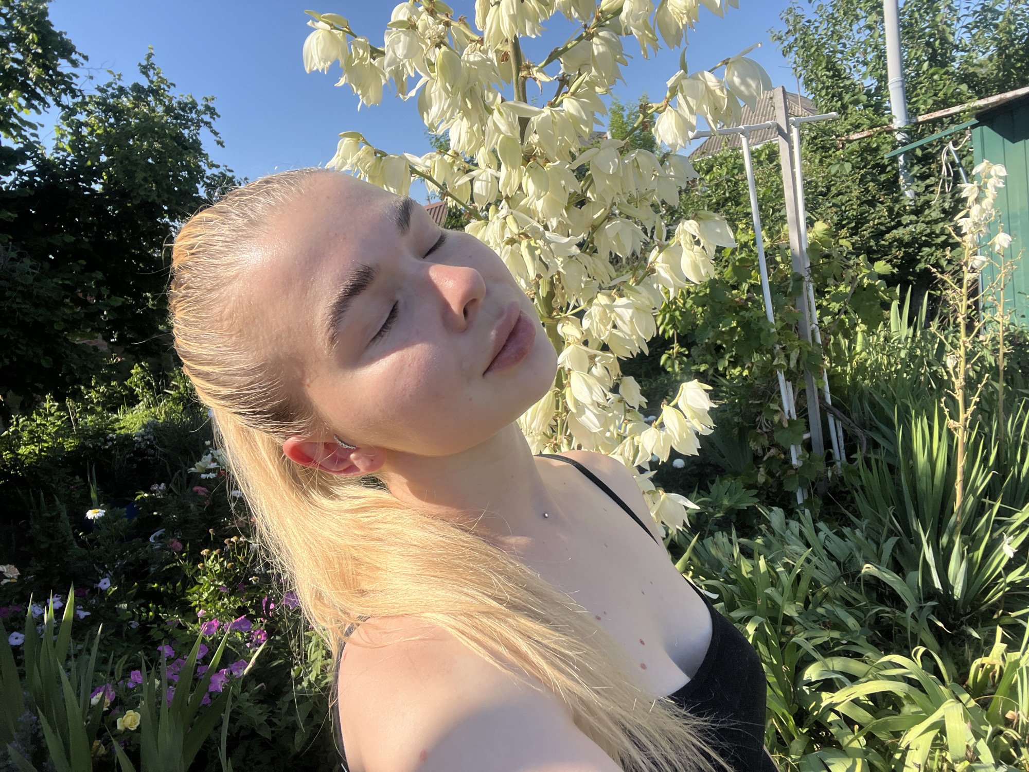 In a sun-drenched garden, a young blonde woman with her eyes closed tilts her head back, basking in the light. Her long, blonde hair cascades over her shoulder. She wears a black tank top, and a small silver stud is visible on her collarbone. The background is filled with lush greenery, including a tall plant with a cluster of white flowers, and a variety of other plants and flowers in bloom, suggesting a temperate climate. In the distance, a glimpse of a rural fence and a weathered building hint at a residential area. The clear blue sky above indicates a fair weather day. The overall impression is one of peaceful enjoyment of nature.