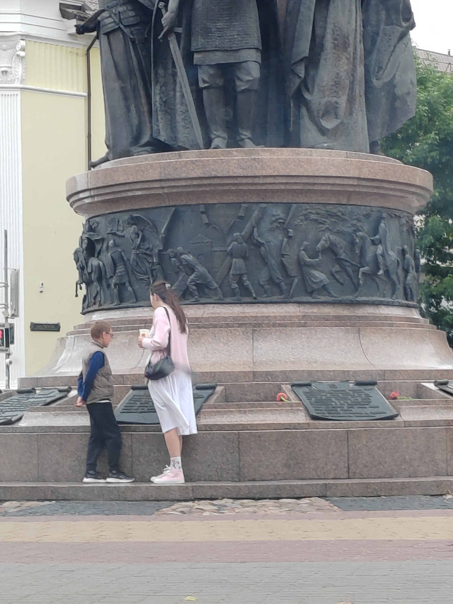 The image shows a low-angle shot of a large bronze monument. In the foreground, two people, a boy and a woman, are standing on a stone base, facing each other. The boy is wearing a vest over a blue shirt and dark pants. The woman is wearing a light pink jacket and a white skirt, holding a cup in her hand. The monument features a detailed bas-relief depicting figures and horses. The monument is located in a city square, with trees and buildings visible in the background. The pavement in the foreground is a mix of grey, yellow, and pink cobblestones. Based on the monument's design, it is likely the Monument to the Millennium of Brest, located in the city of Brest, Belarus.