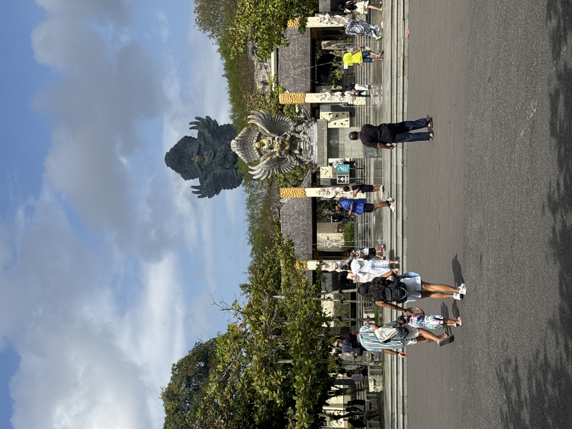 This image captures a sunny day at a monument with a large, dark statue of a mythical creature with wings, possibly Garuda, dominating the left side of the frame. The statue is set against a bright blue sky with scattered white clouds. In front of the statue, there are several people walking on a paved area. The scene appears to be a public attraction or temple complex.
The people are dressed in casual, summer clothing, suggesting warm weather. Some are wearing masks, indicating a concern for health or local regulations. The architecture visible in the background features stone carvings and decorative elements, consistent with a cultural or religious site. The time of day is likely midday, given the strong sunlight and shadows cast on the pavement.
Several individuals are seen walking, some in groups, and others individually. A man in a black shirt and dark pants is standing near the center, looking towards the statue. Further down the path, a group of people are gathered, with some children present. The overall atmosphere seems relaxed and tourist-oriented. The location cues, such as the architecture and the presence of a prominent statue, suggest a place of cultural significance in Kuta Selatan, Indonesia. No discernible text is visible in the image.