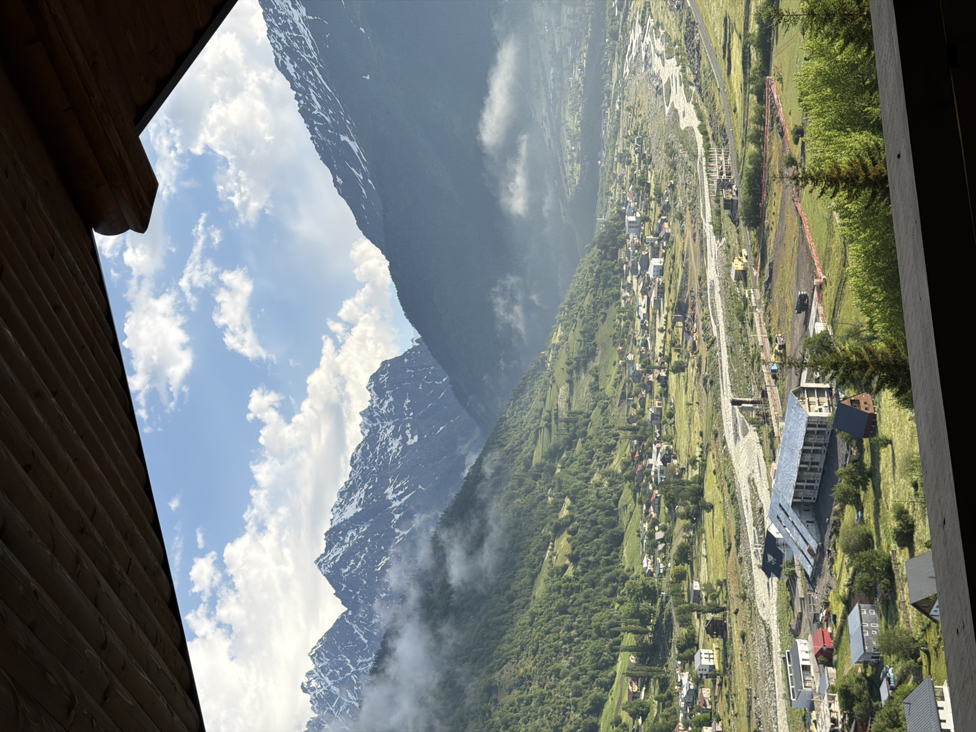 An elevated, outward-facing, landscape-oriented photograph captures a serene view of Mestia, Georgia, situated amidst a valley framed by majestic, snow-capped mountains. The foreground is dominated by a wooden balcony, its dark planks creating a framing effect that partially obscures the view. The sky above is a vibrant blue, dotted with fluffy white clouds that cast soft shadows on the landscape.
The middle ground reveals a sprawling village with numerous houses, their roofs a mix of red and gray, clustered together. A winding, light-colored river carves its way through the valley, bordered by green fields and scattered trees. A modern, elongated building with a metal roof is a prominent feature in this section, alongside other smaller structures and what appears to be a construction site with orange fencing.
The background is dominated by the imposing presence of the Caucasus Mountains. Their lower slopes are carpeted with lush green forests, interspersed with patches of grassland. Higher up, the peaks are adorned with snow, their rugged contours softened by a gentle mist that hugs the mountainsides. The overall scene conveys a sense of tranquility and natural beauty. No people or animals are visible in the frame.