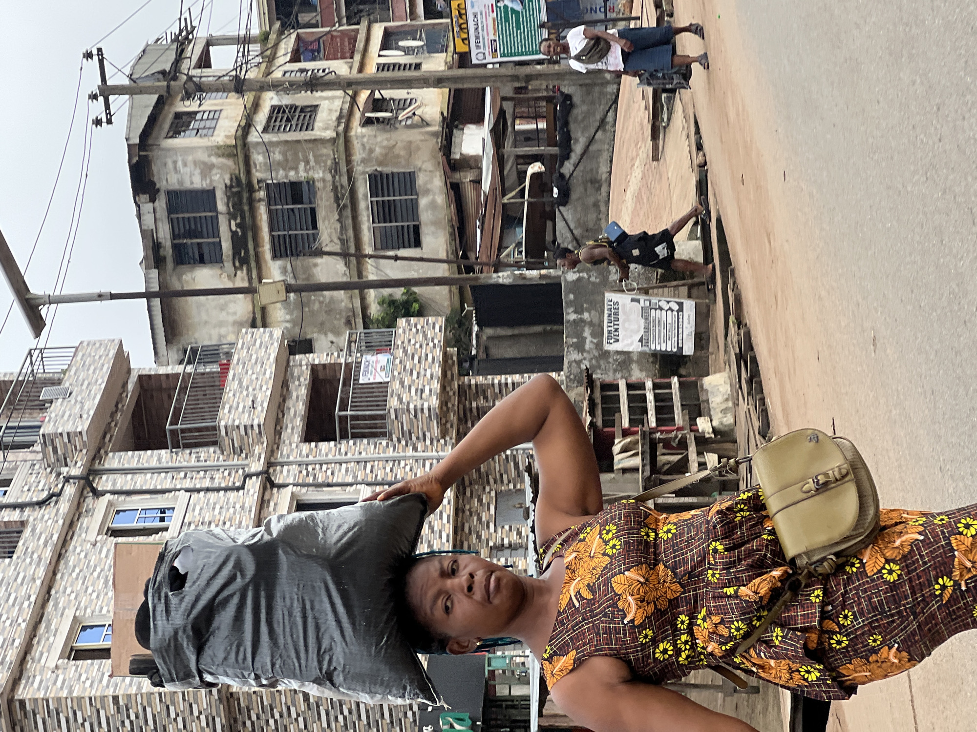 An outdoor, medium shot captures a Black woman in Nkpor, Nigeria, balancing a large, black trash bag on her head. She is walking on a dusty, light-brown road, facing the camera with a neutral expression. Her dark hair is pulled back. She wears a sleeveless, brown dress with a yellow and orange floral pattern, and a green crossbody bag is slung over her left shoulder. In the background, a modern, multi-story building with a brick facade and balconies stands next to older, weathered concrete buildings. The street features a utility pole with wires, various signs, and other people walking in the distance. The sky is overcast.