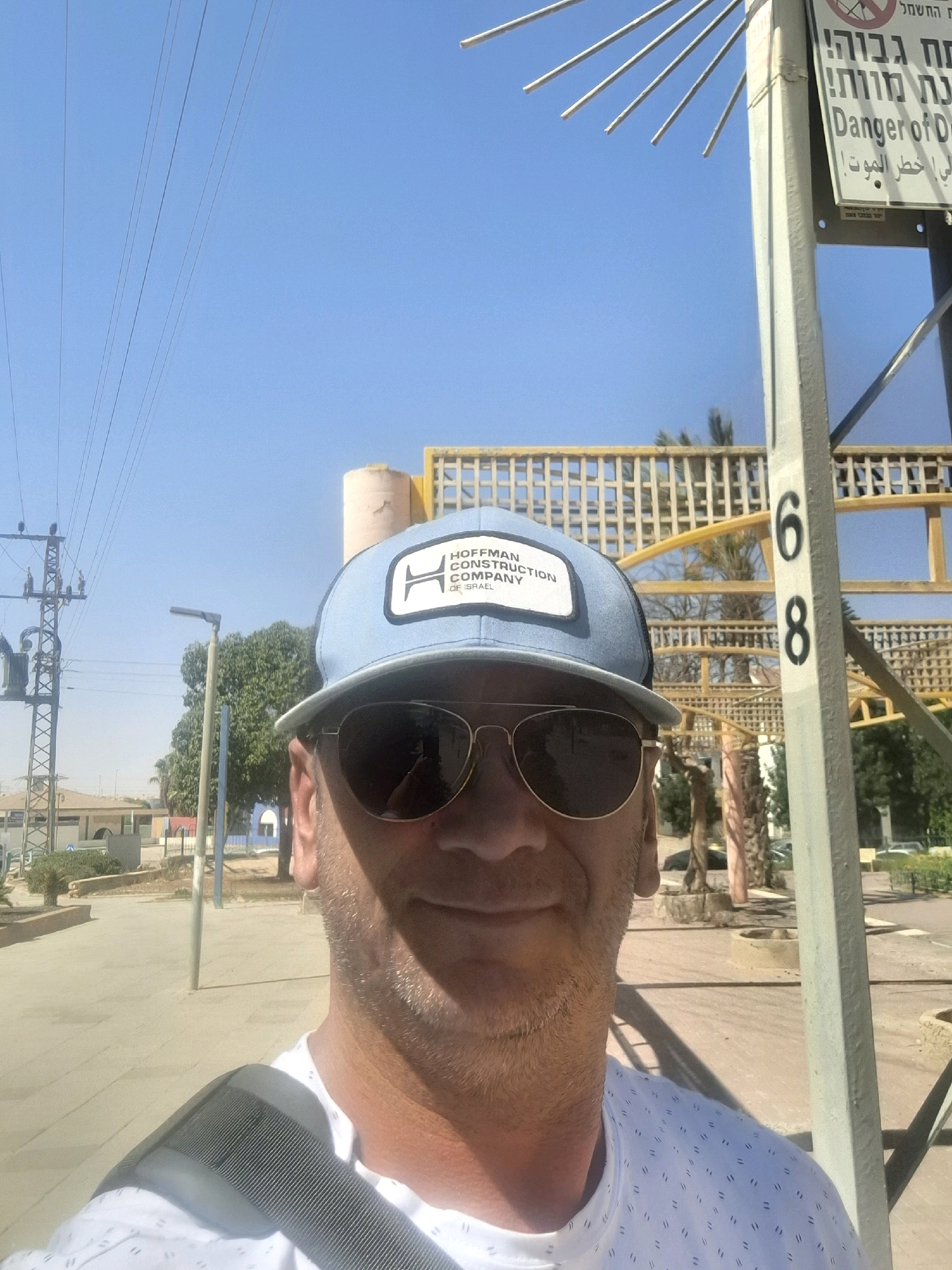 A man wearing a light blue baseball cap and sunglasses takes a selfie outdoors. The cap has a white patch with black text that appears to say "HOFFMAN CONSTRUCTION COMPANY OF ISRAEL." He is also wearing a white t-shirt with a subtle repeating pattern. In the background, there is a clear blue sky with several electrical wires. To the right, a metal pole has the numbers "6" and "8" printed vertically in black. A sign is attached to the pole with text in Hebrew and English, reading "DANGER OF DE..." and "ח ב ר ל מ ו ת!". Further in the background, there are palm trees and some yellow structures. The ground appears to be paved. The overall setting suggests a location in Dimona, Israel, possibly near an industrial or construction area given the signage and structures.