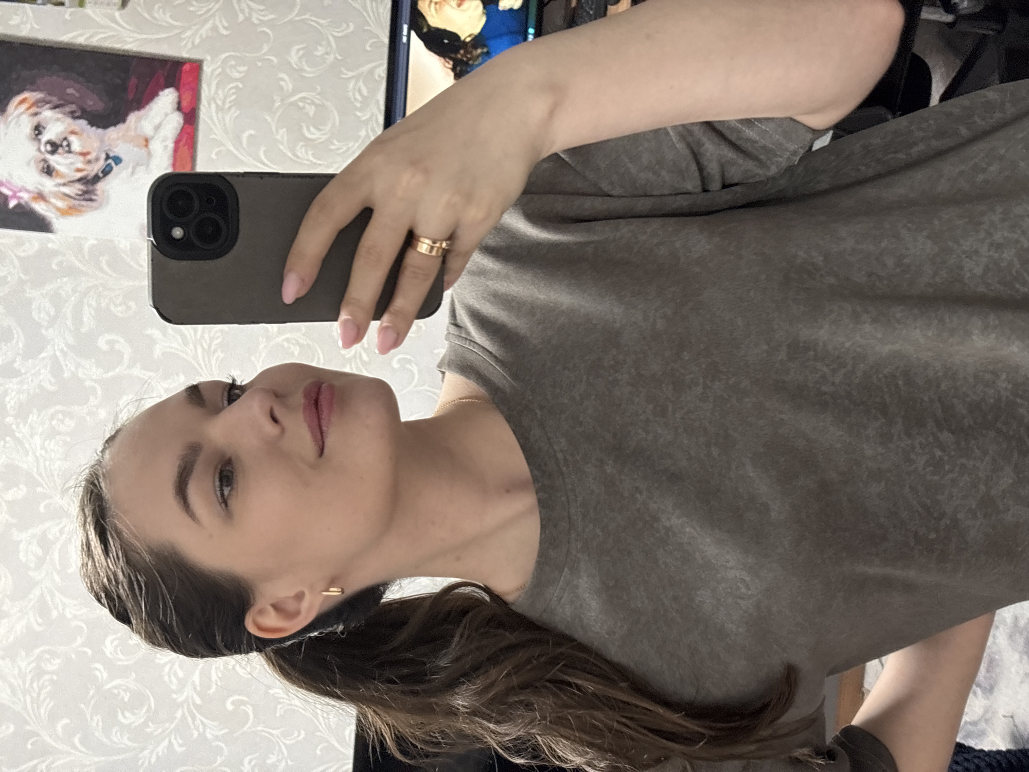 A woman with long brown hair pulled back in a ponytail is taking a selfie in what appears to be an indoor setting. She is wearing a heather grey t-shirt and gold rings on her hand holding a smartphone with a dark grey case. In the background, there is a patterned wallpaper, a picture of a dog, and the edge of a television screen. The exact location within the city of Chaikovsky, Russia, cannot be determined from the image.