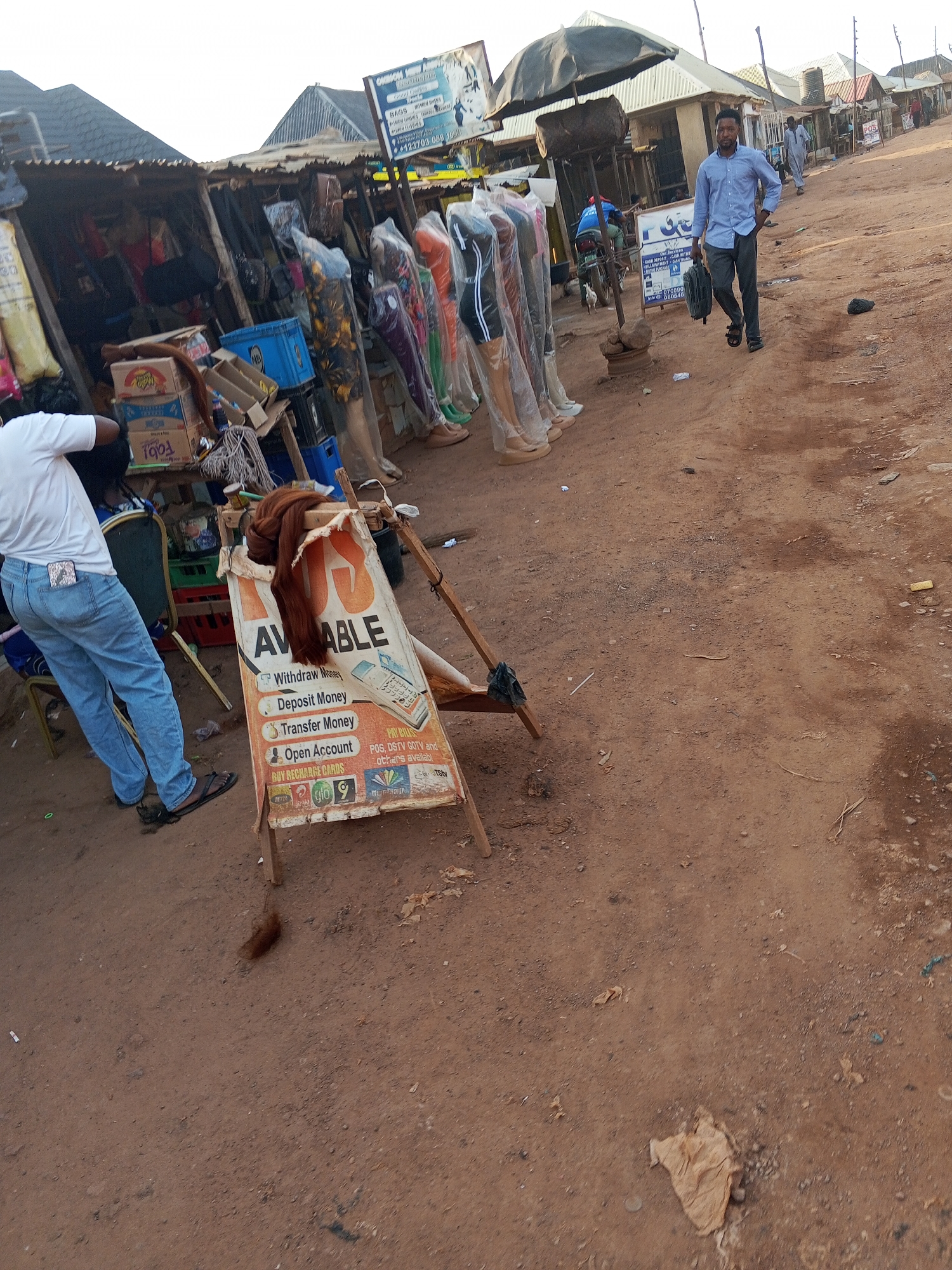 The image shows a street scene in Abuja, Nigeria, characterized by a dirt road and informal commercial activities. A man in a blue shirt and dark trousers walks along the road carrying a bag. To the left, there's a makeshift stall with mannequins displaying clothing, covered in plastic. A sign advertising money transfer services (Withdraw, Deposit, Transfer, Open Account, Pay Bills) is prominently displayed in front of the stall. The stall's supplies are a mix of goods and empty crates. A man stands near the stall, facing away from the camera. In the background, there are other informal shops and structures.