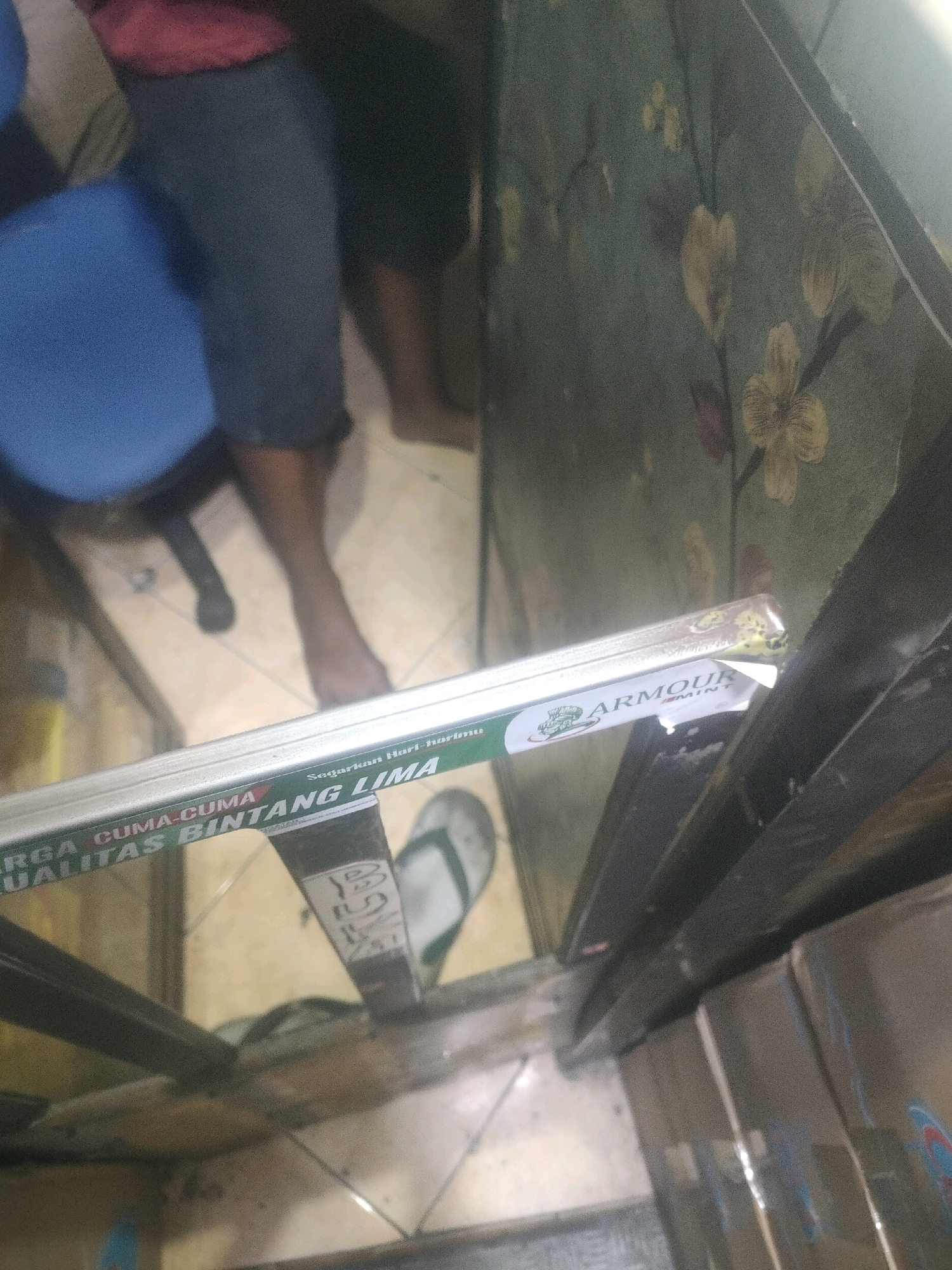 The image depicts a scene within what appears to be a retail environment in Surabaya, Indonesia. In the foreground, a clear plastic package containing what looks like a product with green and white labeling is visible, with text reading "ARGA CUMA-CUMA KUALITAS BINTANG LIMA" and "Segarkan Hari-harimu" and "ARMOUR". The packaging is resting on a metallic shelf or structure. Behind the product, blurred figures of people are present. One individual is partially visible wearing dark blue shorts and a red shirt, and another person is seen with their legs in dark shorts. The floor appears to be tiled. In the background, a wall with a floral wallpaper pattern is discernible. The overall lighting suggests an indoor setting, possibly a shop or market stall.