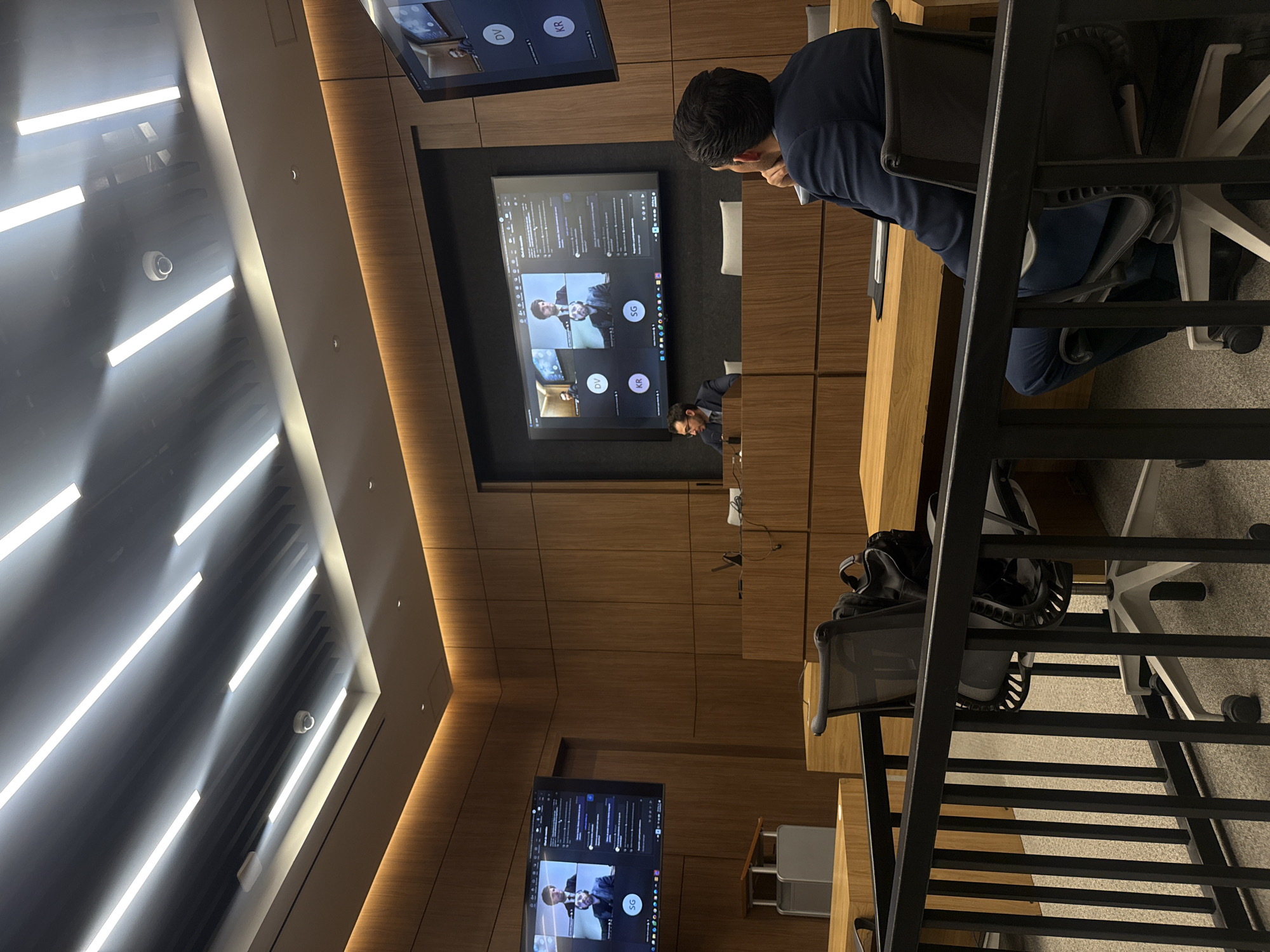 The image depicts a room with wood-paneled walls, likely a conference or lecture hall, in Mexico City, Mexico. On the left side of the frame, there is a wall with several parallel strip lights. A security camera is mounted near the lights. On the opposite side is a wooden desk with a person wearing a dark blue shirt sitting at the desk. Several screens are visible, displaying video conference calls. There are other desks, chairs, and computer gear throughout the room. The scene appears to be set up for a virtual meeting or lecture, with individuals connecting remotely.
