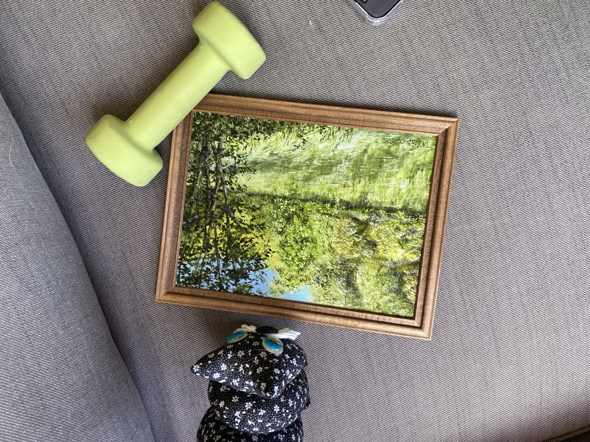 An overhead shot displays an assortment of personal items arranged on a textured grey fabric surface, possibly upholstery, exhibiting diagonal woven lines. Centrally positioned is a rectangular painting enclosed in a light brown, slightly ornate wooden frame. The painting depicts a verdant landscape, dominated by varying shades of green foliage and trees, with a small patch of blue sky visible in the upper left. The lower right corner of the painting bears a signature and date, "M. Kaska 2020." To the left of the painting, a black and white patterned plush item, possibly an animal figure, with prominent light blue "eyes" and white tufts, rests on the fabric. Above and to the right of the painting lies a lime green, short dumbbell. In the bottom right corner of the frame, a segment of a smartphone encased in clear plastic is partially visible.