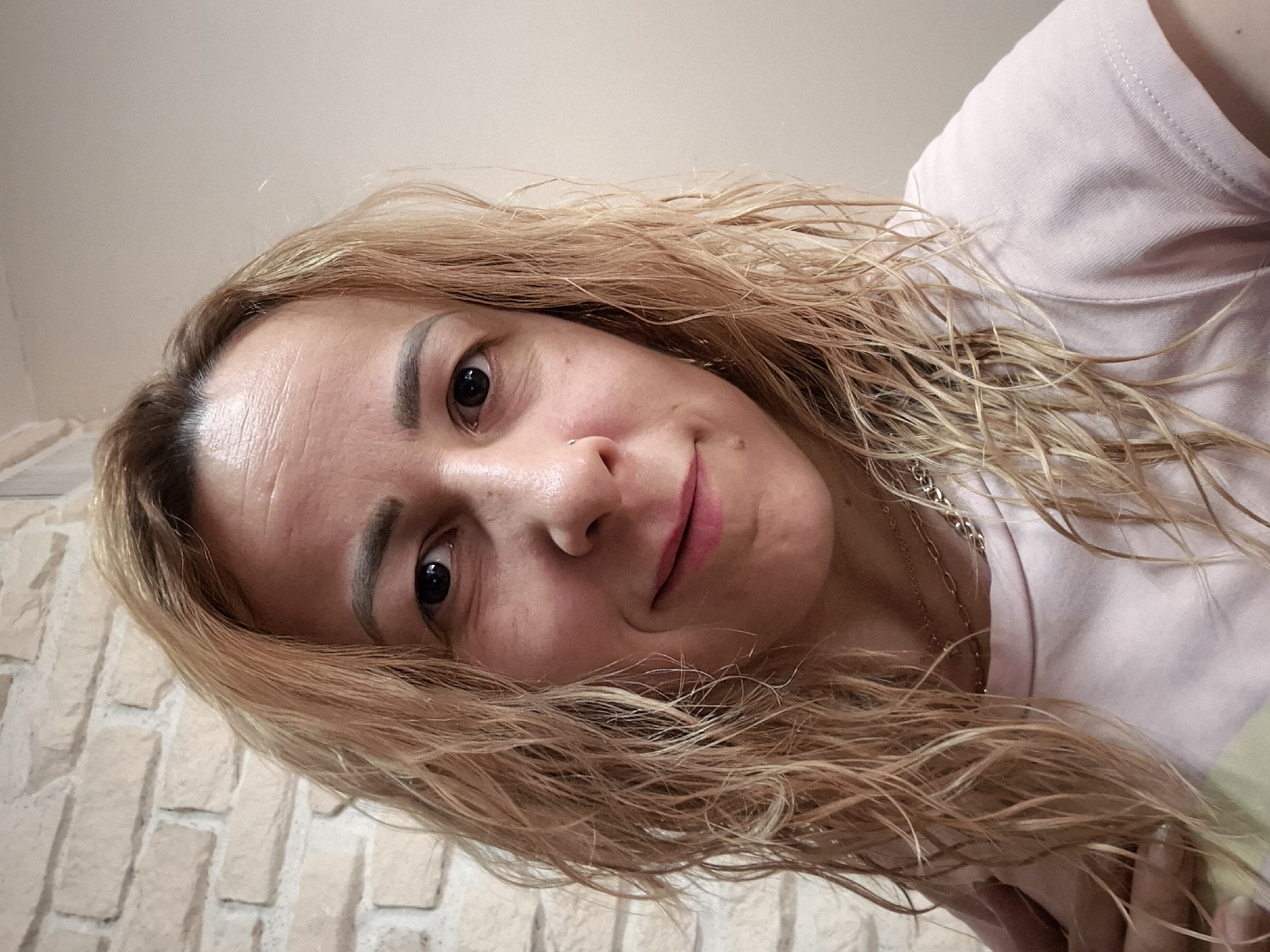 A woman with light blonde, wavy hair is positioned with her head tilted downward towards the camera. She appears to be wearing a light pink shirt and a necklace is visible around her neck. A brick wall is partially visible in the lower left corner of the image. The location is not identifiable as within the city of Unknown, Unknown.
