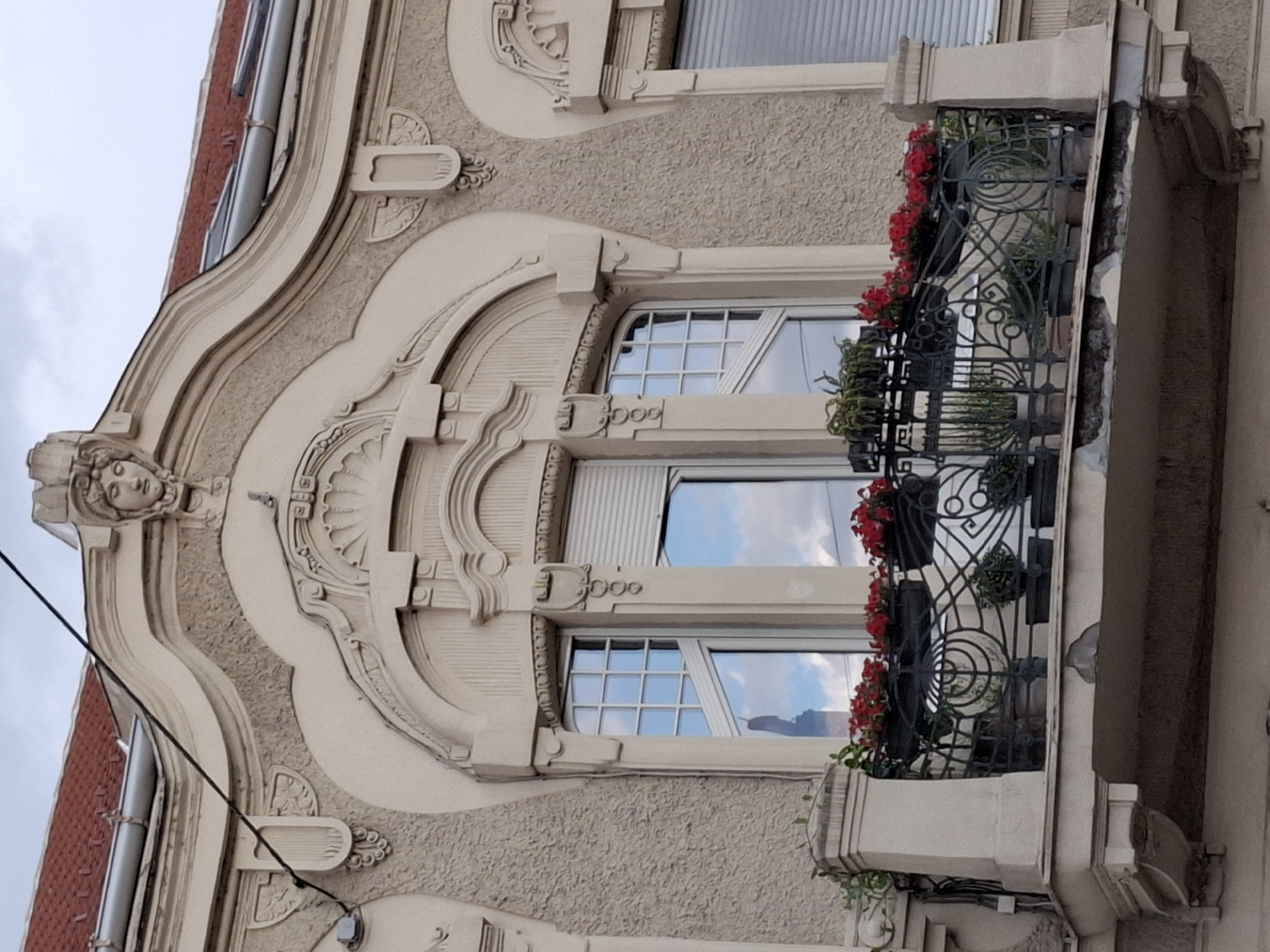 The image shows the facade of a building with ornate architectural details, including a decorative plasterwork arch above a window, a bust of a woman at the apex of the arch, and a balcony with wrought iron railings. The balcony is adorned with potted plants, including red flowers. The building is rendered in a textured stucco material. The sky is overcast, with reflections of clouds visible in the window panes. The scene is located in Zemun Urban Municipality, Serbia, with the architecture suggesting a style common in that region. No people are visible in the image.