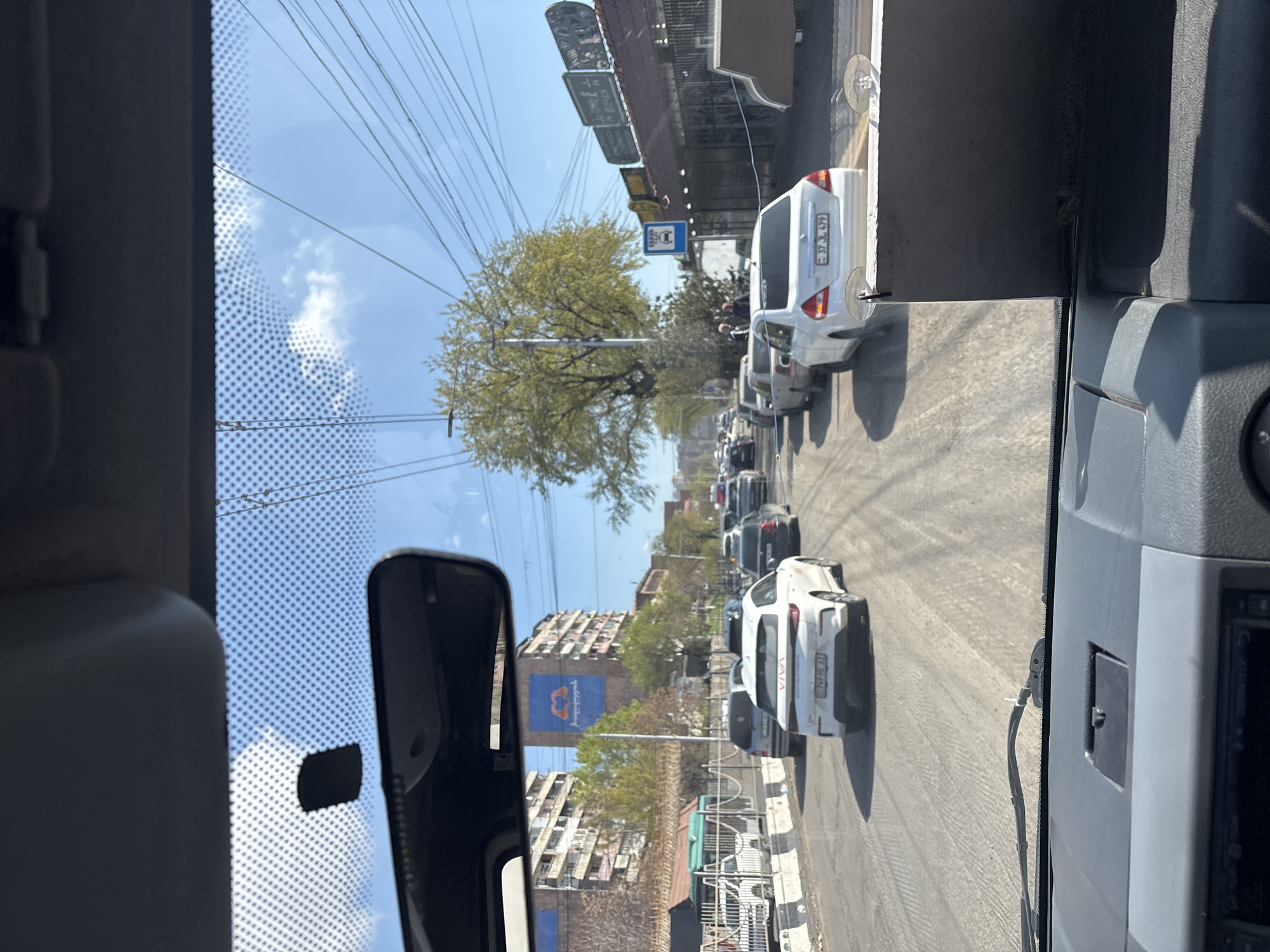 A view from inside a vehicle, likely a car or bus, looks out onto a city street. Traffic is congested, with multiple cars lined up on the road. A large tree is visible in the middle ground, and buildings are seen in the background, one of which features a large blue advertisement. Overhead, numerous power lines crisscross the sky. The scene appears to be a typical urban environment. The license plate reads "37 JJ 479" which corresponds to Yerevan, Armenia.