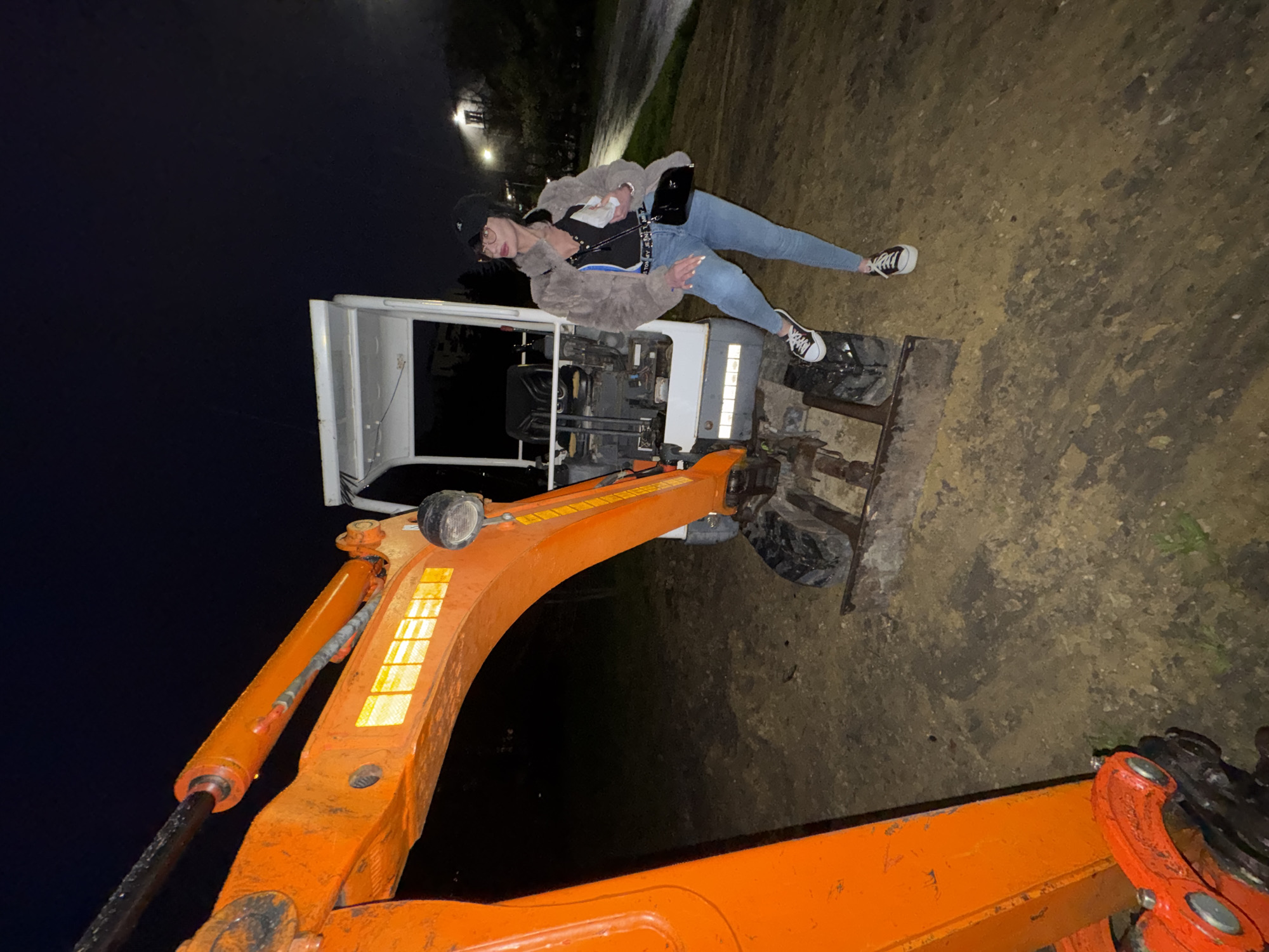 The image displays a woman posing on an orange and white mini excavator at night in an outdoor, unpaved setting. The woman is positioned on her back across the engine section of the excavator, with her legs extended to the side. She wears light blue jeans, black and white sneakers, a black top, a fluffy grey jacket, a black baseball cap, and glasses. A black shoulder bag is slung across her.
The excavator is predominantly orange, featuring a white cabin structure and visible hydraulic components, some adorned with yellow reflective tape. The ground surrounding the machinery is dark and appears to be dirt or gravel, possibly muddy or damp. The scene is illuminated by artificial light, creating highlights on the subjects and equipment while the background remains largely dark. Faint lights are visible in the distant background, suggesting a nearby building or street. No visible text is discernible in the image. The photograph captures a casual, possibly playful moment in Varna, Bulgaria, with industrial equipment as an unconventional prop.