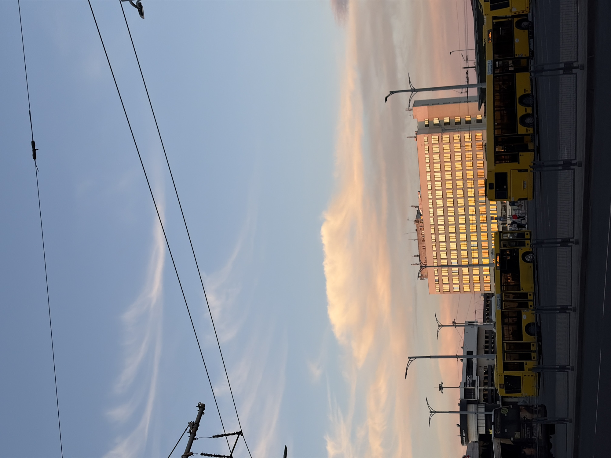 The image, originally rotated 90 degrees clockwise, depicts an urban scene in Minsk, Belarus, at either sunrise or sunset. The sky dominates the upper portion, a clear light blue with delicate, wispy clouds stretching across, tinged with soft pink and orange hues from the low sun.
In the midground to the right, a large, multi-story building stands prominently. Its numerous rectangular windows brightly reflect the golden light of the sun, casting a warm glow across its facade. Below and to the right of this building, a row of long, yellow trolleybuses is parked along a paved street or dedicated bus lane. Their wheels are visible, and some appear to have passengers inside, though indistinct. Overhead, an intricate network of dark power lines, characteristic of trolleybus systems, spans across the sky, connected to utility poles on the left and right sides of the frame. Streetlights with curved arms are also visible, some extending above the parked vehicles.
On one of the yellow buses, the route number "63" is clearly visible, along with faint Cyrillic text that includes "СЕРЕБРЯНКА" (Serebryanka), a residential district in Minsk. Another bus displays "КОНКОРО" (Konkoro), possibly a company name or advertisement. Further text, "ТЦ" (TC - Shopping Center), is faintly discernible on a lower building.
The overall atmosphere is calm and serene, capturing the transition of day to night or night to day in a bustling city, with the warm light providing a gentle contrast to the otherwise functional urban infrastructure. The activity appears to be routine urban transit, with buses either waiting or having completed their routes.