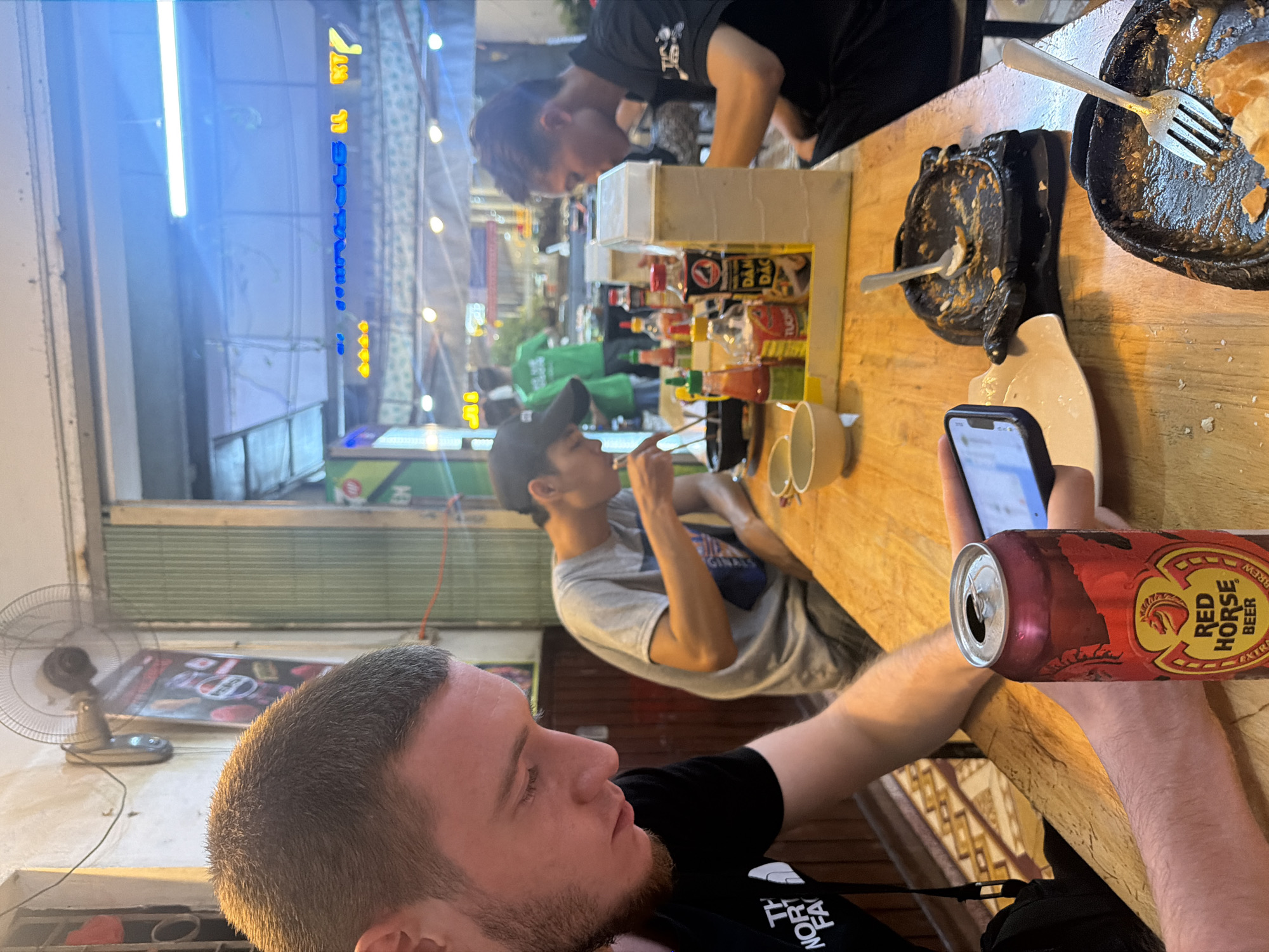 This image depicts a casual eating establishment in Nha Trang Ward, Vietnam, likely an open-front restaurant or street food stall. In the foreground, a Caucasian male, appearing to be in his 20s-30s with short brown hair and a beard, is seated at a wooden table, wearing a black "THE NORTH FACE" t-shirt. He is looking down at a smartphone held in his right hand. An open red and silver can of Red Horse Beer is positioned on the table directly in front of him.
In the mid-ground, an Asian male wearing a grey t-shirt and a dark baseball cap is seated, leaning forward, and eating from a bowl with chopsticks. Behind him and to the right, another Asian male with dark hair tied back, wearing a black polo shirt, is also seated and appears to be eating from a bowl.
The wooden table holds several items: two black, possibly cast-iron, sizzling plates with remnants of food and cutlery (a spoon in one, a fork in the other); multiple small, light-colored ceramic bowls; and a yellow plastic caddy containing various Vietnamese sauces, including chili sauce ("TƯƠNG ỚT"), dark soy sauce ("TƯƠNG ĐEN"), and a bottle labeled "DẬM ĐẶC".
The background reveals a simple, functional environment. The wall to the left is a light-colored, unadorned surface with a wall-mounted electric fan and some exposed wiring. Through an open entrance to the right, a bustling street scene is visible with various illuminated signs, including blue and yellow neon text (e.g., "KT7"), and string lights overhead. Corrugated metal shutters are partially visible on the right side of the entrance. Further back, two individuals in green shirts, presumably staff, are partially visible, with one standing with his back to the camera. The floor beneath the table features a light-colored tiled pattern.