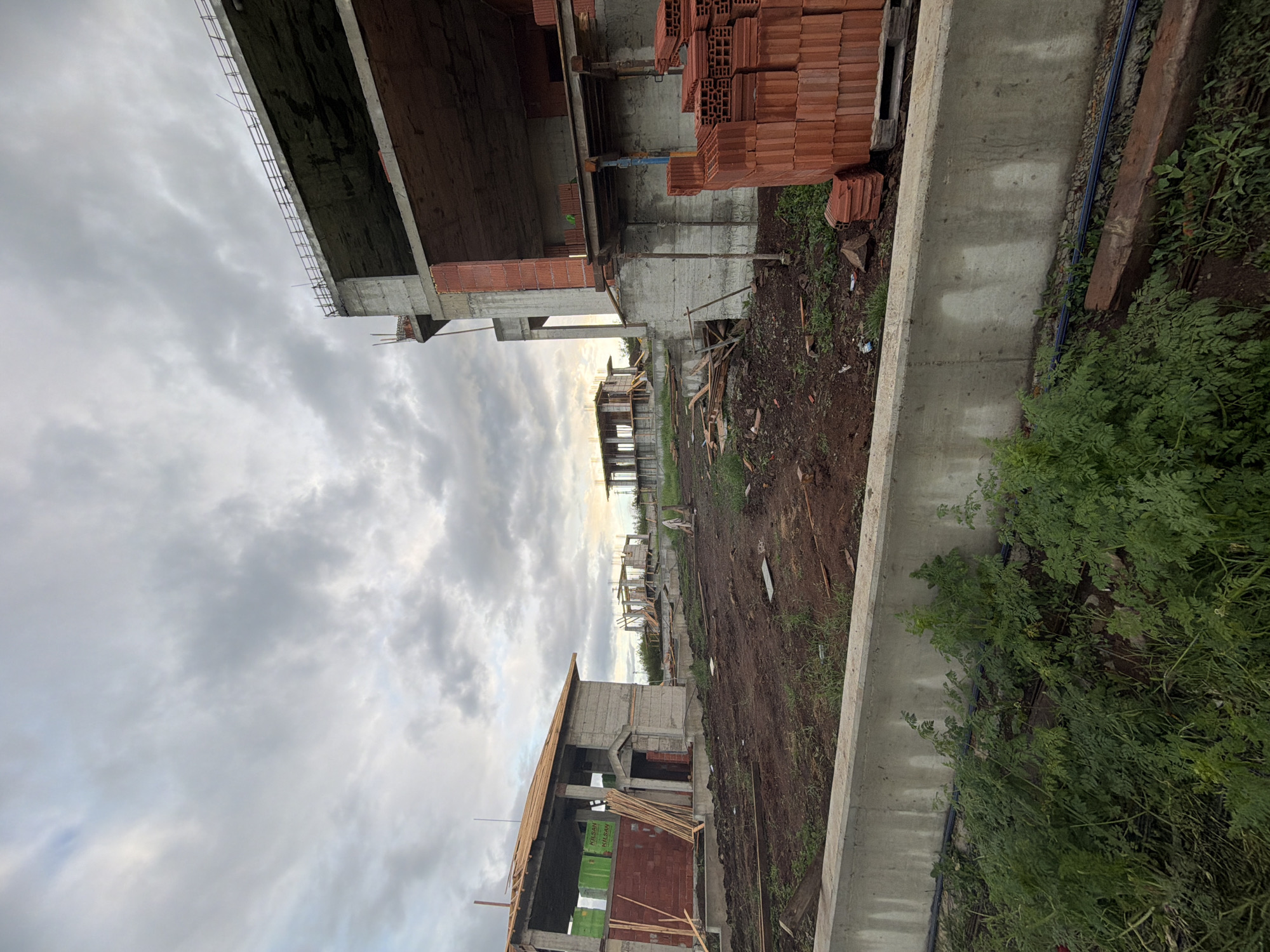 The image depicts a construction site in Cherni Vrah, Bulgaria, with unfinished buildings of reinforced concrete and brick. Stacks of bricks are visible near one of the structures. A concrete retaining wall runs along the edge of the site, separating it from an area with dense green vegetation. The ground is muddy and disturbed, typical of construction activity. Overcast skies fill the upper portion of the image.