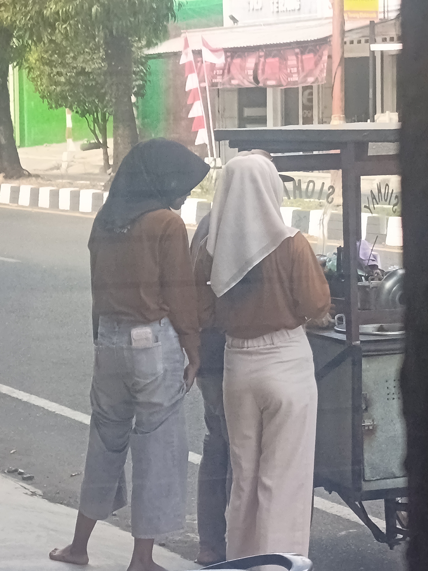 The image captures a street scene in Desa Gunungsimping, Indonesia, focusing on two individuals standing near a food cart. The person on the left, wearing a dark hijab and a brown long-sleeved shirt, has light-colored pants and a light-colored item in their back pocket. The person on the right wears a light-colored hijab and a brown long-sleeved shirt, paired with light-colored trousers. They are positioned next to a food cart, which appears to be a mobile vendor. The background shows a street with a pedestrian walkway, a road with a white line marking, trees with green foliage, and buildings. The overall lighting suggests it is daytime.