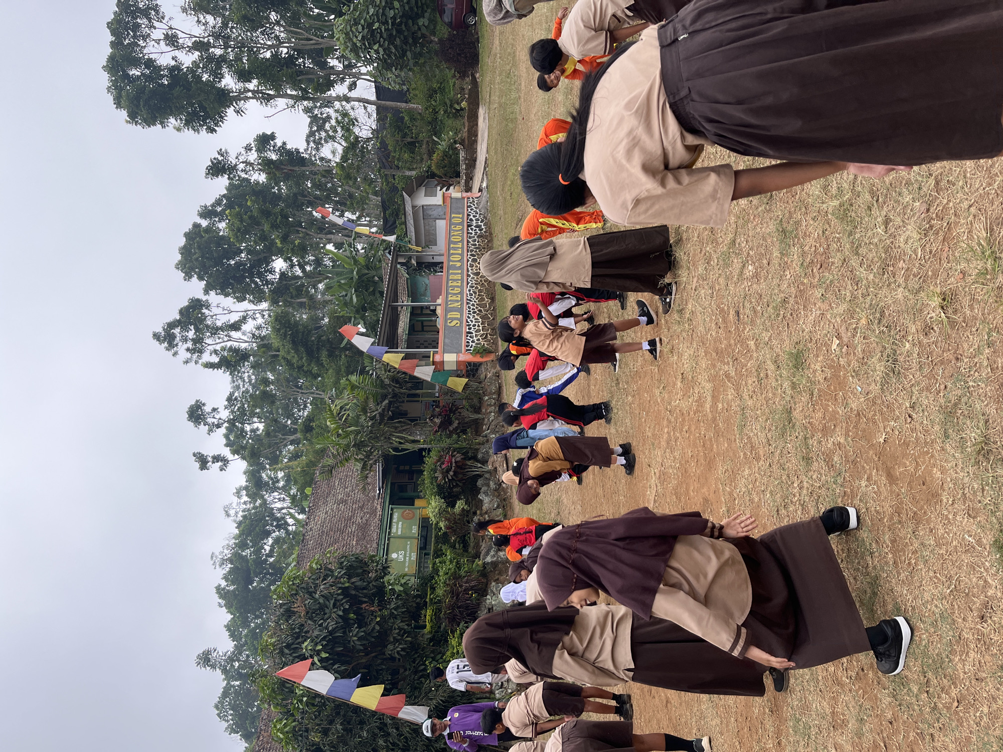 The image depicts an outdoor scene at SD Negeri Jollong 01, a primary school in Gembong, Indonesia. A group of students, predominantly wearing uniforms consisting of light brown tops and dark brown skirts or trousers, are engaged in what appears to be a physical activity or exercise session on a dirt and grass field. Some students are bending forward, while others are standing or in motion. A few individuals are wearing headscarves. In the background, a modest school building with a tiled roof is visible, along with trees and foliage. Indonesian flags are displayed on poles. The sky is overcast.