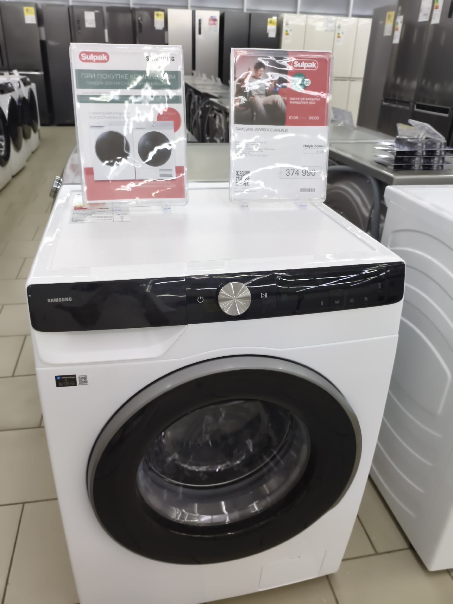 In a retail environment, likely a large electronics or appliance store, a white Samsung washing machine is prominently displayed. It has a black control panel with a silver dial and several buttons. A black door with a glass window reveals the drum inside. Two promotional signs are attached to the front of the washing machine. The sign on the left, with a red and white design, promotes a discount on dryers when purchasing a washing machine. The sign on the right, featuring an image of a person on a couch and a red circular logo, advertises a bonus for a purchase with a start and end date. The floor is tiled in a light color. In the background, rows of other appliances, primarily refrigerators, are visible. The setting is indoors and well-lit, characteristic of a commercial establishment. The image appears to be taken in Aktobe, Kazakhstan, based on the presence of Cyrillic text on the promotional materials.