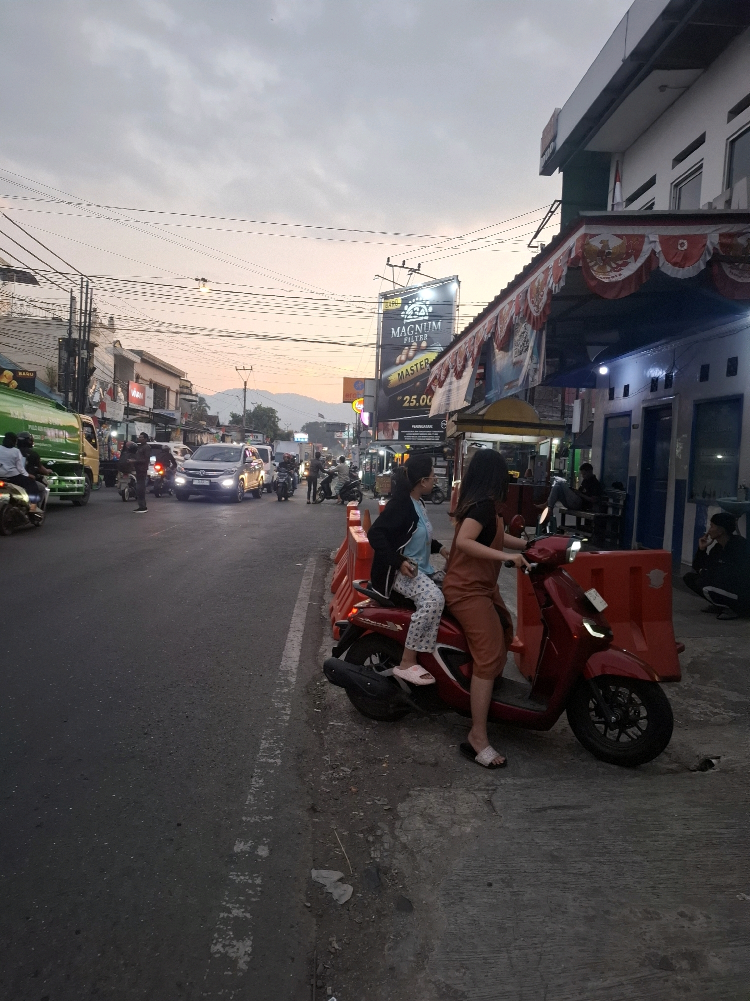 The image depicts a street scene in Cikaroya, Indonesia, during the early evening. The sky is overcast with a gradient of pale orange and grey, suggesting either sunrise or sunset. Several vehicles are visible on the road, including a silver SUV in the foreground with its headlights on, a green truck on the left, and numerous motorcycles. Two women are seated on a red scooter in the middle of the frame; the woman in the back is wearing a black jacket and patterned pants, while the woman in front is wearing an orange jumpsuit and a black top. They are positioned in front of a building with an awning decorated with Indonesian flags. Several orange traffic barriers are placed along the side of the road. A billboard for "MAGNUM FILTER" is visible in the background, along with various storefronts and utility poles with a dense network of wires. The overall atmosphere is that of a busy urban street with ongoing traffic and commercial activity.