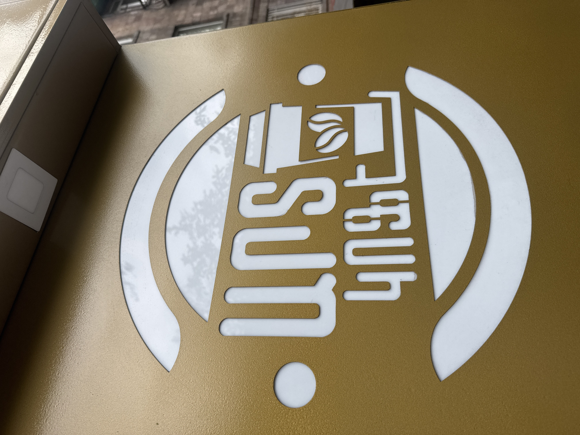 The image shows a close-up of a sign for "SUT", presumably a business establishment. The sign features a stylized logo in white against a gold-colored background. The logo depicts a coffee cup with coffee beans inside, with the name "SUT" written below, and a symbol resembling Wi-Fi waves enclosing the design. Based on previous knowledge, this sign is located in the city of Yerevan, Armenia. There are no people visible in the image. The environment appears to be outdoors, based on the building features at the top of the shot.