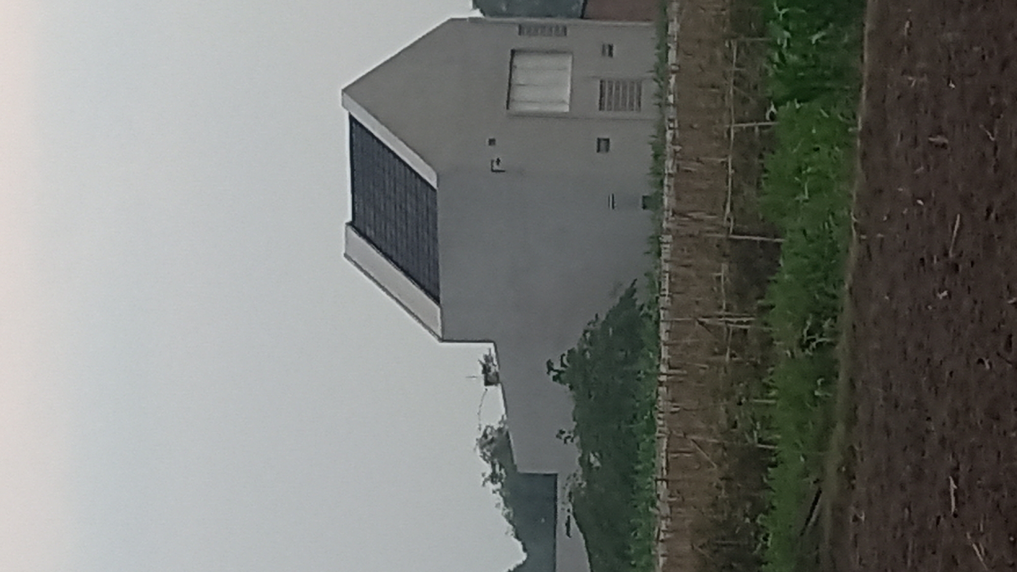 A modern, minimalist house with a grey, stucco exterior and a dark, sloped roof is visible in the middle ground. The house has large, rectangular windows on its front facade. The surrounding environment consists of agricultural fields, with rows of dry vegetation and green crops in the foreground and middle ground. The sky is overcast and grey. The scene appears to be located in a rural or semi-rural area. The image is taken from a distance, and the resolution is moderate. No people are visible in the image.