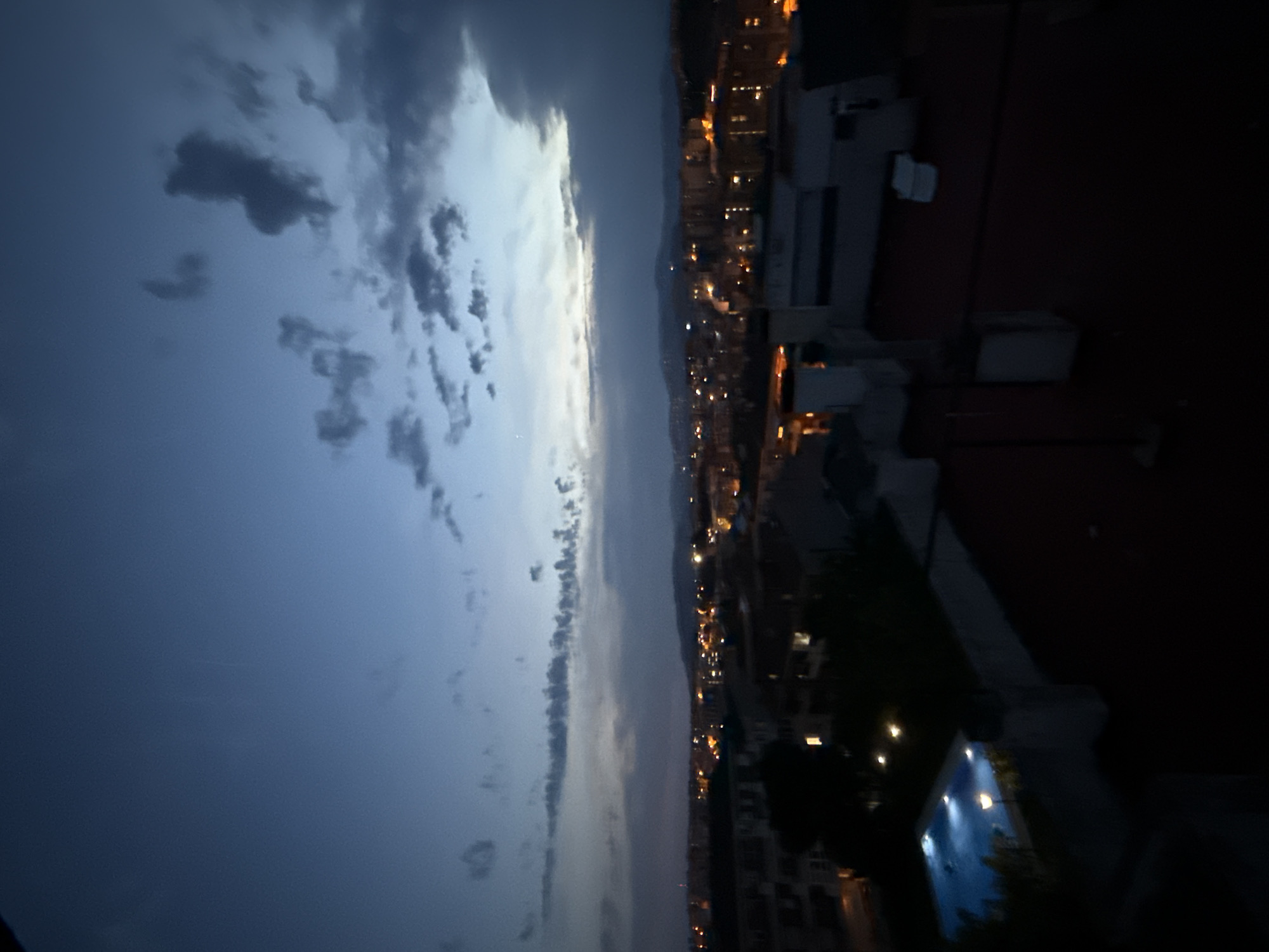 This is an exterior, eye-level, wide shot of a cityscape at dusk. The sky is a deep, twilight blue, with scattered, dark clouds. In the foreground, a dark rooftop with red tiles is visible. To the left, a brightly lit blue swimming pool is visible with several illuminated lights under the water. Beyond the rooftop, the city of Blanes, Spain, stretches into the distance, with buildings illuminated by numerous orange and yellow lights. Hills can be seen in the background.
