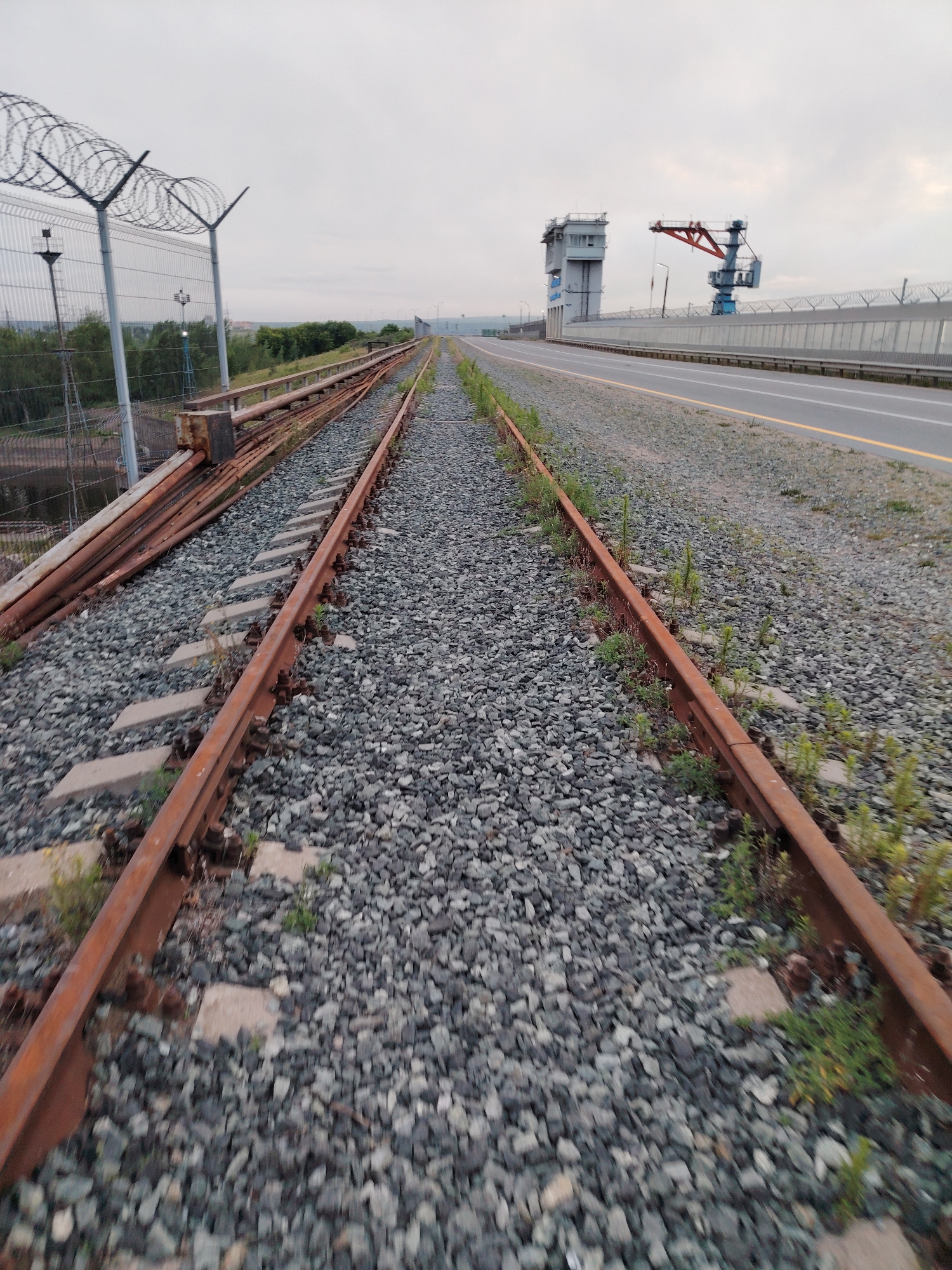 An outdoor, eye-level, vertical, slightly blurred, long-shot photograph captures an abandoned railroad track stretching into the distance. The track is composed of rusty metal rails and is covered in gray gravel. On the left side, a chain-link fence with barbed wire runs parallel to the tracks. Beyond the fence, trees and some industrial structures are visible. On the right side of the tracks, a paved road with a yellow line runs alongside. In the background, a large, gray, multi-story building with windows is situated next to a blue and red construction crane. The sky is overcast and a pale gray. The location is the city of Chaikovsky, Russia, specifically an area near the Chaikovsky Hydroelectric Power Station.