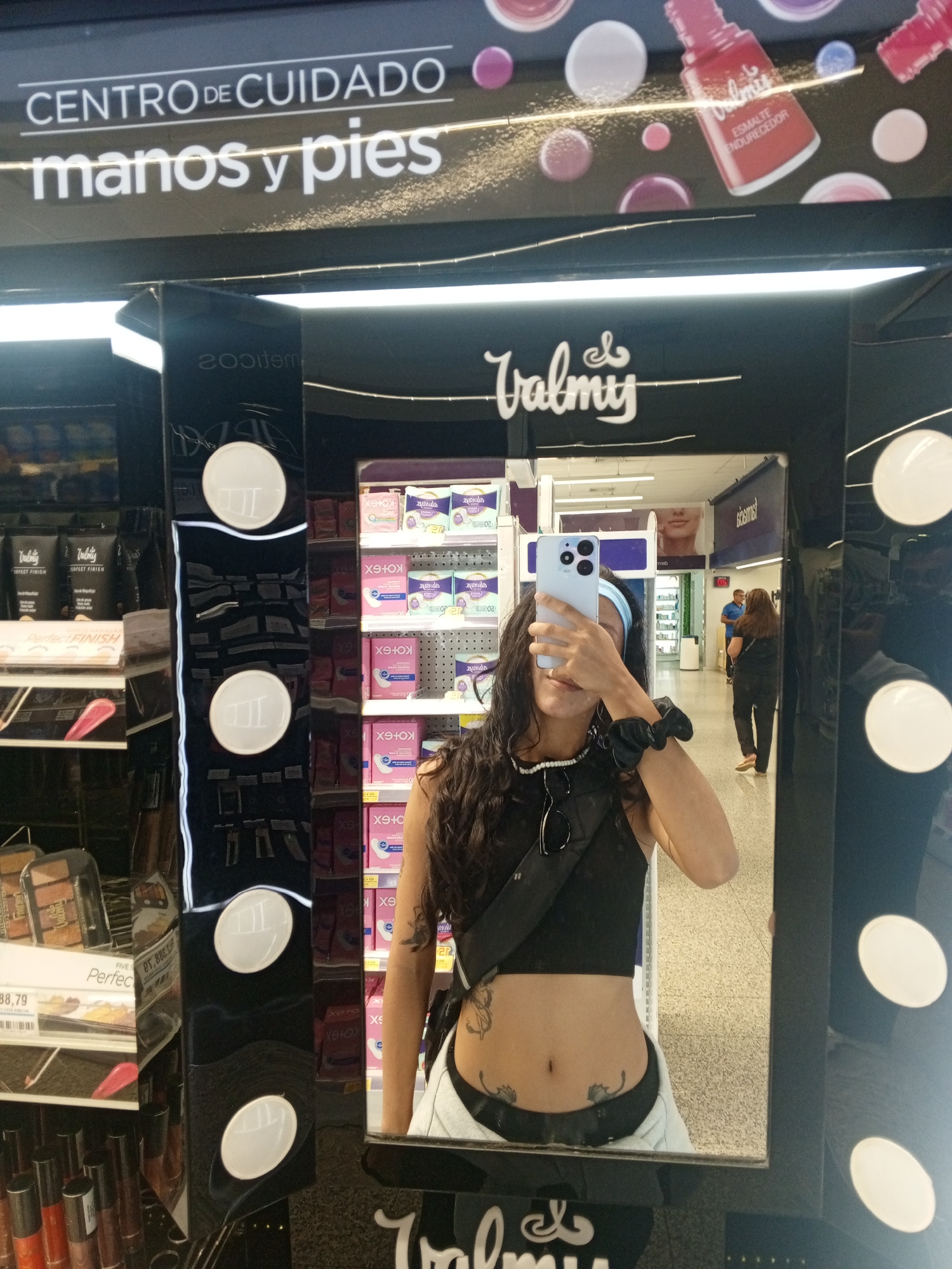 A woman is taking a selfie in a mirror in a store in Pampatar, Venezuela. The mirror is part of a Valmy brand display, featuring nail polish and other cosmetics. The woman has dark hair, a black crop top, tattoos on her abdomen, and is wearing a light blue headband. The store sells Kotex feminine hygiene products. Other shoppers are visible in the background. The words "CENTRO DE CUIDADO manos y pies" appear above the display.