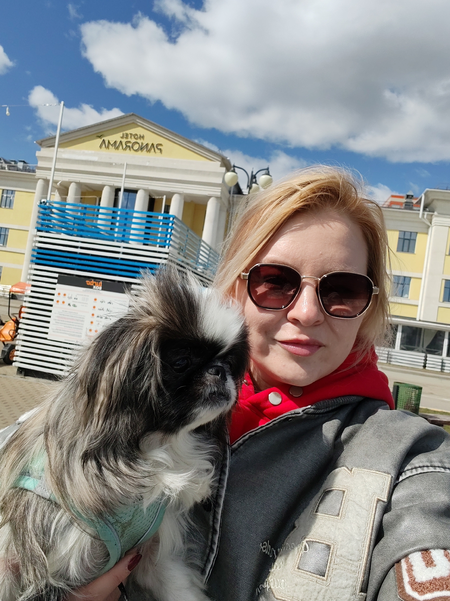 A medium shot depicts a woman holding a dog in front of the Hotel "Amaks Central" in Izhevsk, Russia.
The woman, light-skinned with blonde hair and wearing sunglasses, is angled slightly to the left, looking into the camera. She is wearing a red hoodie underneath a gray leather-like jacket with patches on the sleeve and shoulder.
The dog, primarily black and white with long fur, is held in her left arm, looking to the left. It is wearing a teal harness.
Behind the woman and the dog is a building identified as the "Amaks Central" hotel, its facade painted yellow and white. A temporary blue and white striped structure is visible in front of the hotel. The sky is blue with scattered white clouds. The shot is taken outdoors in daylight. The ground appears to be a paved surface. An orange electric golf cart is visible.