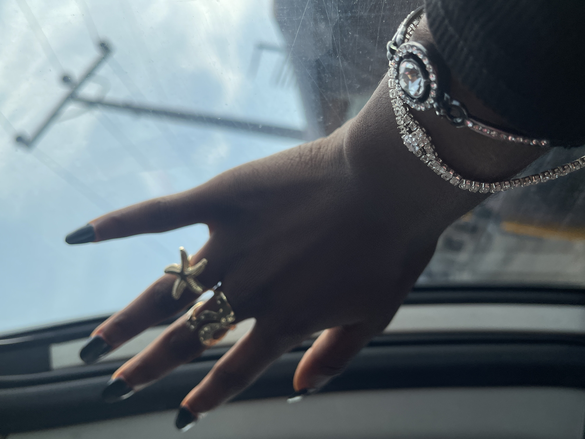 A close-up shot shows a dark-skinned hand adorned with jewelry. The hand has long, dark-polished nails, and the fingers are decorated with two gold rings: one resembling a starfish on the index finger and another appearing as a serpent on the middle finger. A jeweled bracelet is worn on the wrist. The hand is positioned in front of a slightly blurred window, which reveals a glimpse of a bright sky and what appears to be an electrical power pole. The scene implies the subject is inside a vehicle, possibly in transit. This image was most likely not taken in Ado, Nigeria. Ado Ekiti is an area in Nigeria, not a city. Ado, Nigeria is also not a city.