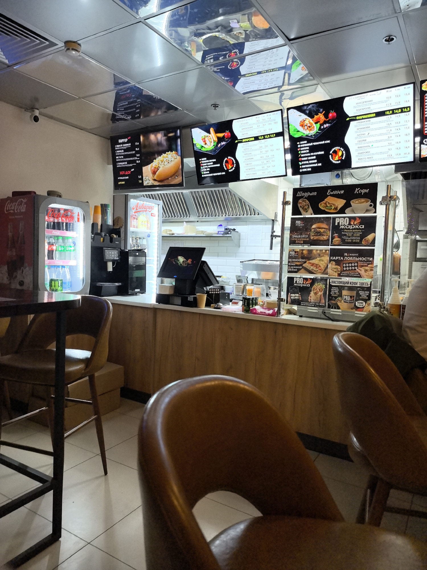The image depicts the interior of a fast-food establishment, possibly a shawarma or kebab shop, with menu boards prominently displayed. In the foreground, two brown leather chairs are partially visible, suggesting a dining area. The counter area features a Coca-Cola branded refrigerator, a drink dispenser, and a POS system. Behind the counter, a kitchen area with stainless steel appliances and an exhaust hood is visible. Digital menu boards display food items and prices in Cyrillic script, with images of various wraps, burgers, and drinks. The overall environment is brightly lit, with a clean and functional design characteristic of a quick-service restaurant. There are no visible people in the image.