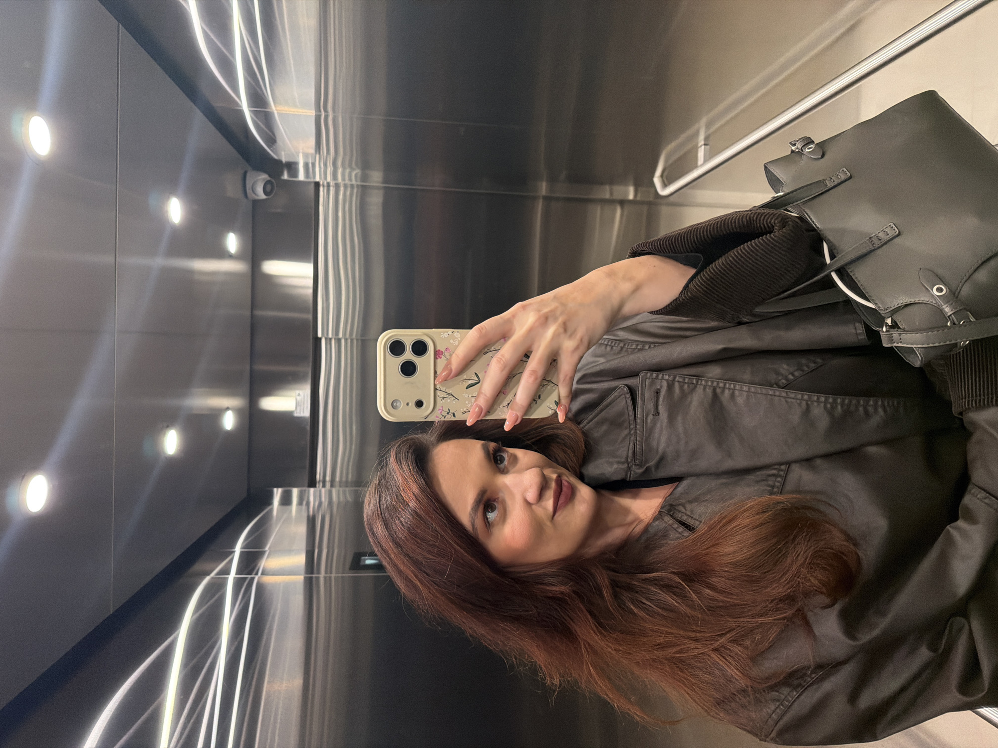 A woman with long, auburn hair and a floral-patterned cell phone case is taking a selfie in what appears to be an elevator. She is wearing a dark jacket. A handbag is next to her. The elevator has polished steel walls, lighting fixtures, and a security camera in the upper left corner. The location appears to be in an elevator somewhere in Belgrade, Serbia, based on visual inspection and general city characteristics.