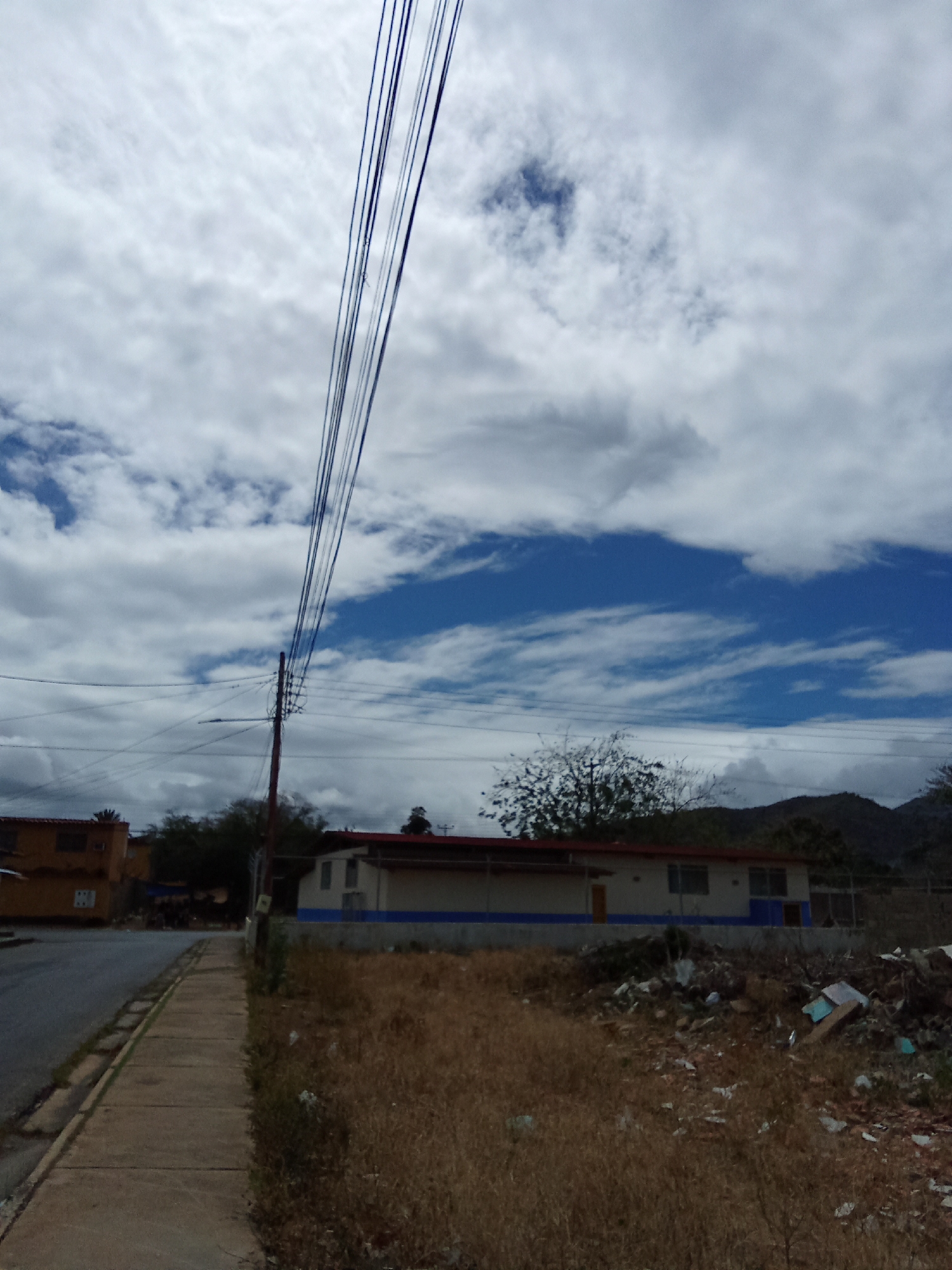 The image presents a street scene in Juan Griego, Venezuela. In the foreground, a sidewalk runs alongside a paved street. A power pole with multiple wires dominates the left side of the image. Adjacent to the sidewalk is an unkempt area filled with dry vegetation and discarded debris, including plastic and cardboard. A low building with a red roof and white walls, trimmed with a blue stripe, is visible in the background. A mountain range is visible further back. The sky is filled with clouds and patches of blue sky. No people are visible.