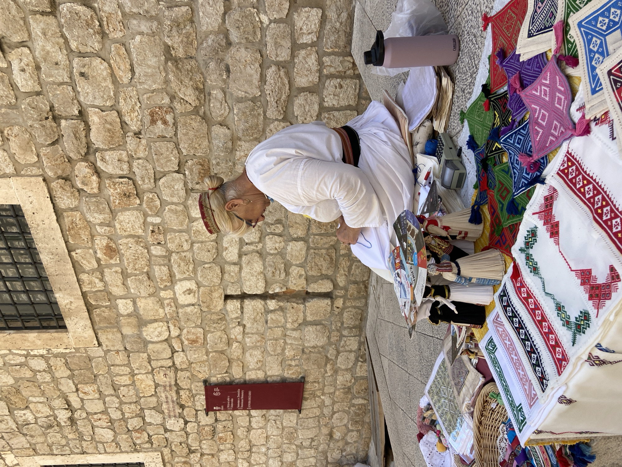A woman in traditional Croatian attire is sitting in front of a stone wall in Dubrovnik, Croatia, displaying her handicrafts. The woman has blonde hair pulled back in a bun and wears a white dress with a dark sash. She is wearing glasses and appears to be sewing or working on one of her crafts. In front of her is a display of various embroidered textiles, including napkins, tablecloths, and small decorative items. There are also dolls dressed in traditional clothing. The stone wall behind her is part of an old building, possibly a historical site or a market stall. A sign in Croatian is hanging on the wall. The scene suggests a cultural and artisanal market, likely in the historic Old Town of Dubrovnik.