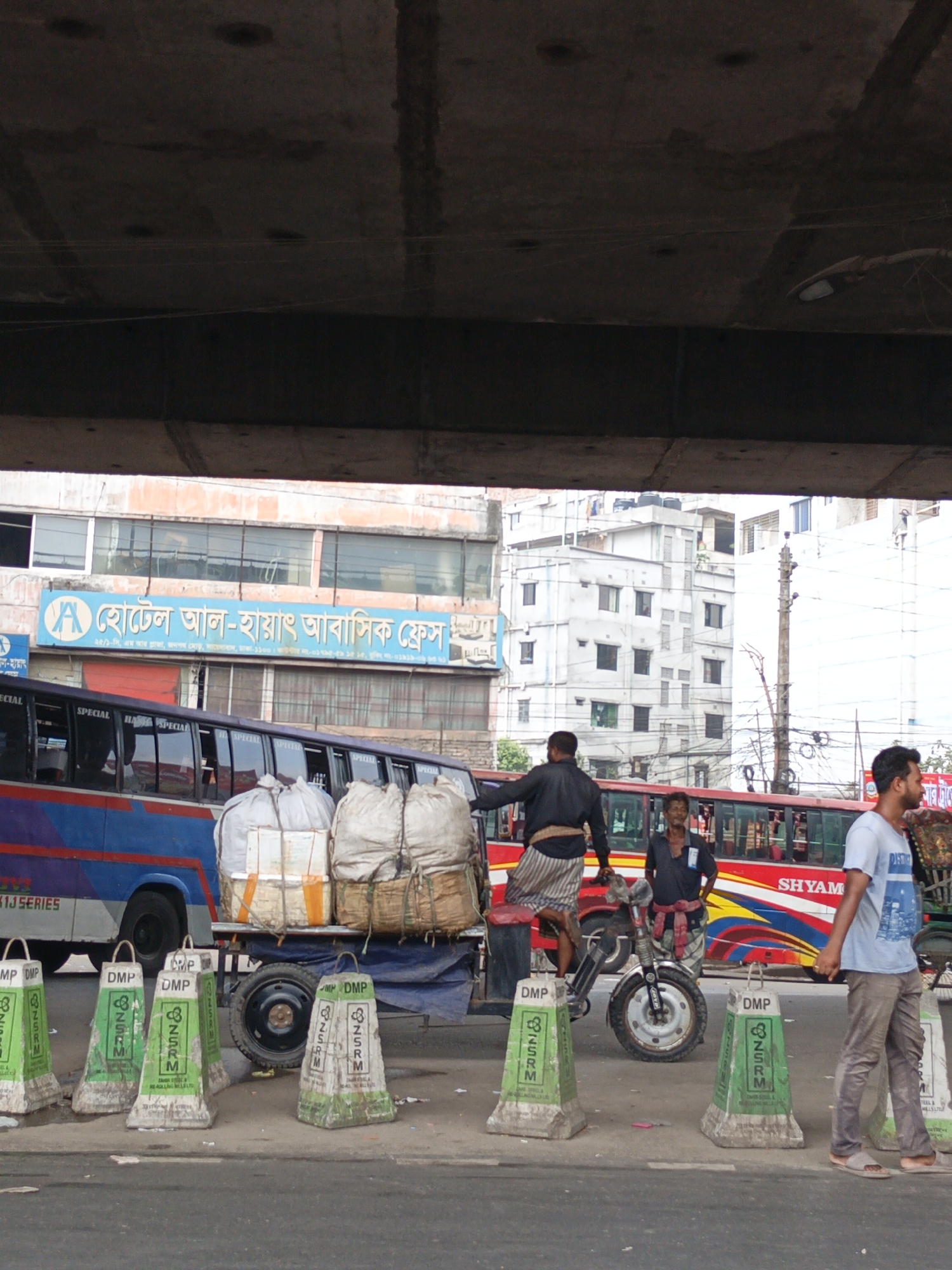 The scene captures a street in Dhaka, Bangladesh, under an overpass. In the foreground, a series of green and gray concrete traffic barriers labeled "DMP" (Dhaka Metropolitan Police) and "NSRM" (a steel manufacturing company) line the street. Behind them, a three-wheeled motorcycle rickshaw is heavily loaded with large, white sacks.
A man wearing a black shirt and striped shorts is standing on the rickshaw. To the right, another man in a dark shirt stands near the rickshaw, while a man in a blue t-shirt and gray pants walks by.
In the background, blue and red passenger buses are visible, indicating this is likely a busy area for public transport. A sign for "Hotel Al-Hayat Abasik Fress" is also visible, suggesting a commercial district. The architecture includes a mix of older and newer buildings, typical of Dhaka's urban landscape. Overhead, the concrete structure of the overpass dominates the upper portion of the image, creating a shadowed effect.